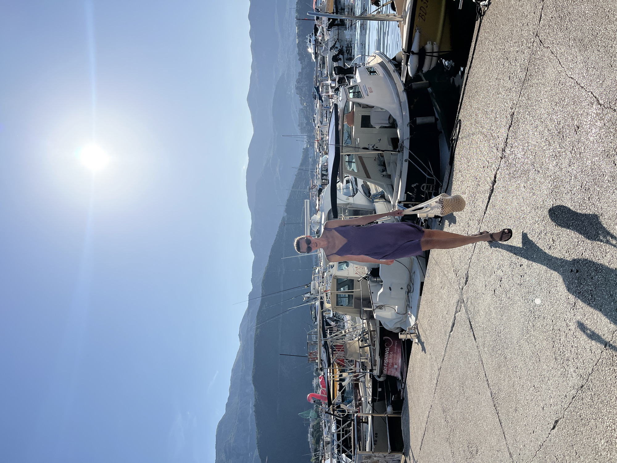 This image captures a woman walking on a sunlit concrete pier in a marina, identified as Budva, Montenegro, by signage on a boat. The woman, positioned in the lower-mid ground, is walking forward and slightly to the viewer's right. She wears a sleeveless purple dress, dark sunglasses, and dark flat sandals, carrying a light-colored woven net bag on her left arm. A long shadow extends behind her, cast by the strong sunlight originating from the upper right of the frame.
The marina is densely filled with various boats, predominantly small to medium-sized motorboats and fishing vessels, docked in calm, reflective water to the woman's right and extending into the background. One boat prominently displays a sign that reads "BOAT-FISHING" with "Budva, Montenegro" visible below it. Another boat features a distinctive large pink inflatable flamingo on its deck. In the background, a substantial range of dark blue-grey mountains spans the horizon, with clusters of buildings visible at their base. The sky above is a clear, bright blue, with the sun appearing as a brilliant light source with a lens flare in the upper right quadrant. The concrete pier surface shows cracks and wear.