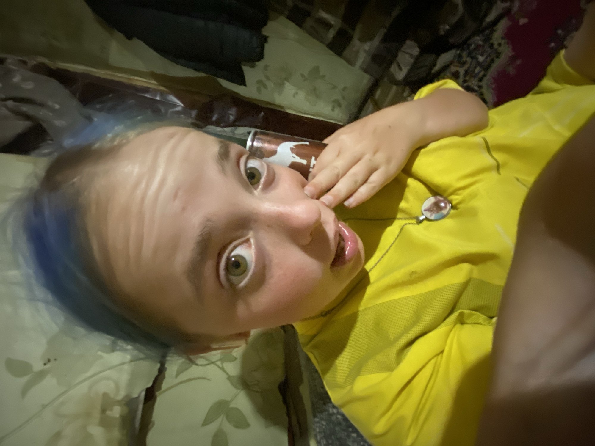 A close-up, low-angle selfie captures a young person with bright blue hair and wide green eyes. They are wearing a vibrant yellow t-shirt and a silver necklace with a circular pendant. Their left hand is raised to their cheek, and their mouth is slightly open, suggesting surprise or curiosity. In the background, there appears to be a patterned fabric, possibly bedding or upholstery, in muted green and brown tones. To the right of the person, a cylindrical object with a white horse logo is visible, possibly a beverage can. The overall setting appears to be indoors, with dim lighting contributing to a somewhat cluttered and informal atmosphere. The specific location within Nova Praha, Ukraine cannot be determined from the provided image.