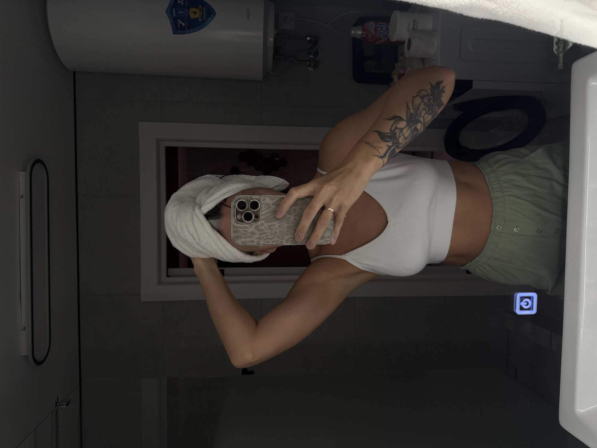 A woman in a bathroom, with a white towel wrapped around her head, is taking a selfie in a mirror. She is wearing a white top and light green shorts. Her left arm has a prominent tattoo. The location appears to be a bathroom in Saratov, Russia, as indicated by the "ZIYO" logo on a water heater affixed to the wall. The background includes standard bathroom fixtures: a toilet, toiletries, and a towel rack.