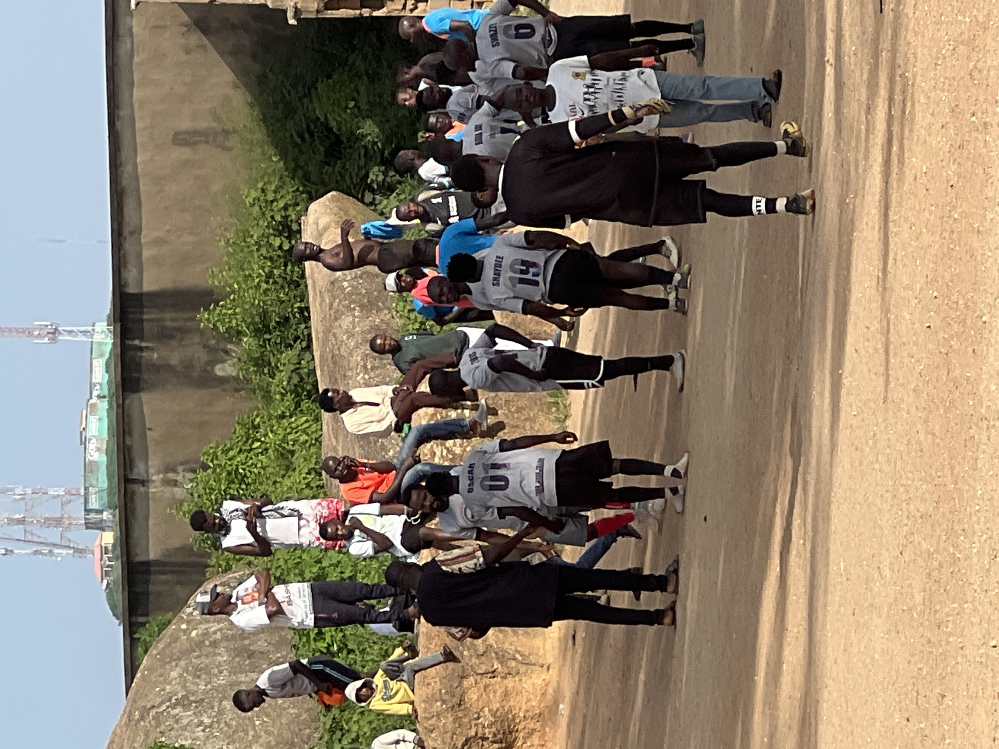 The image depicts a group of men gathered outdoors on a dirt ground, possibly during a sporting event or informal meeting, in Jos, Nigeria. The setting appears to be a rugged area with large rock formations and a concrete wall in the background. A few individuals are wearing athletic attire, including jerseys with numbers and names such as "OSCAR 01" and "SHAYDEE 19". Some men are standing, while others are seated on the rocks. One person is holding a mobile phone, and another appears to be engaged in conversation. The environment is sunny, suggesting daytime. The overall impression is a candid moment captured in a community setting.
