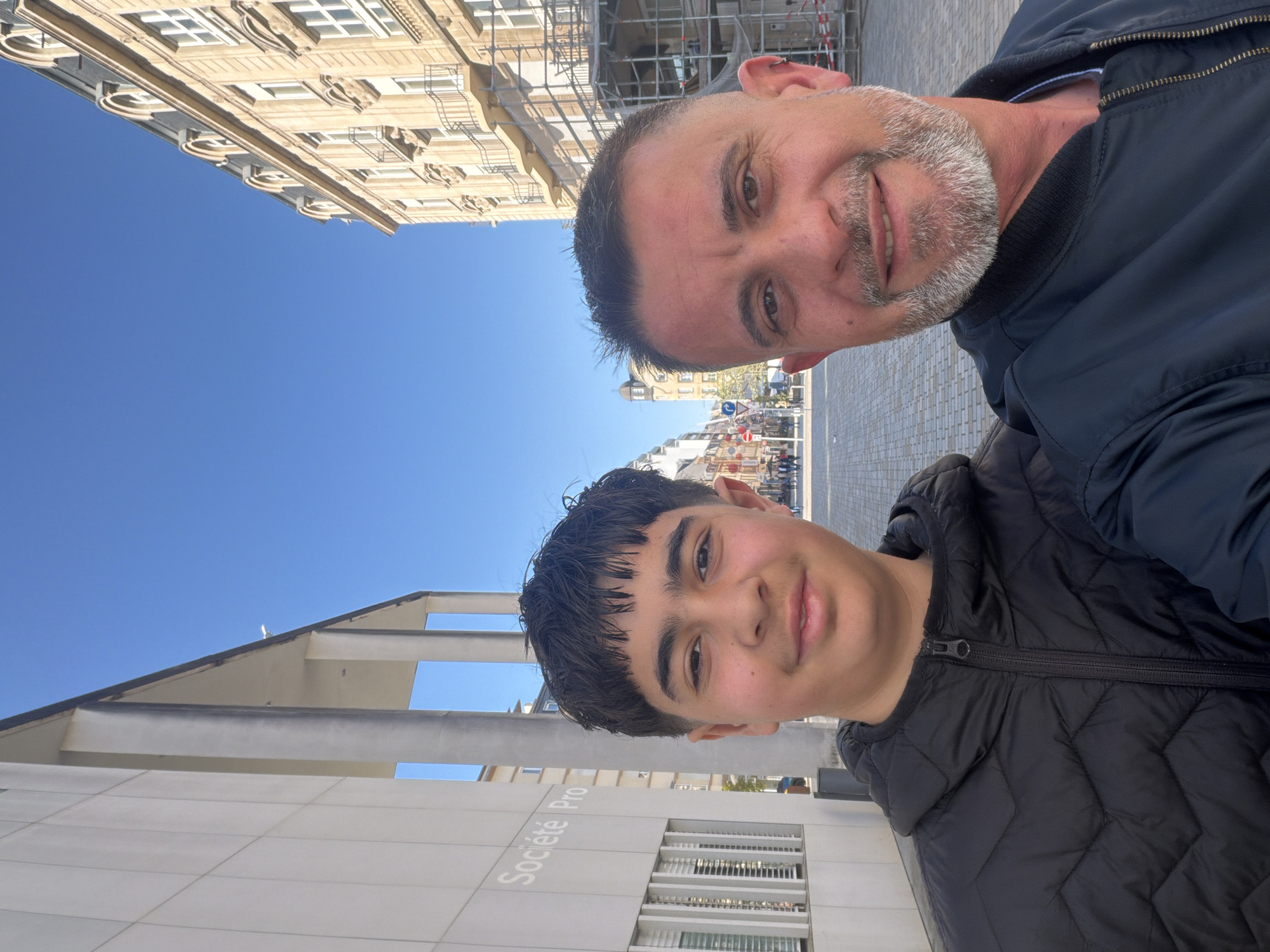 A selfie captures two males outdoors in Esch-sur-Alzette, Luxembourg, on a clear, sunny day. The man on the right, older with short dark hair and a grey beard, wears a dark bomber-style jacket and smiles warmly. Beside him, a younger individual with dark hair and a fringe also smiles, wearing a black quilted hooded jacket.
The background reveals an urban environment. To the left, a modern building with a light-colored facade features the text "Société Pro" printed vertically. Its architecture includes angular elements and large windows. To the right, a classic European-style building with ornate detailing, stone or stucco finish, and several balconies is partially covered by scaffolding, suggesting construction or renovation. Beyond these buildings, a street or paved plaza is visible with additional buildings and street signs in the distance. The sky above is a bright, cloudless blue, indicating pleasant weather. Both individuals appear content and happy as they pose for the camera.