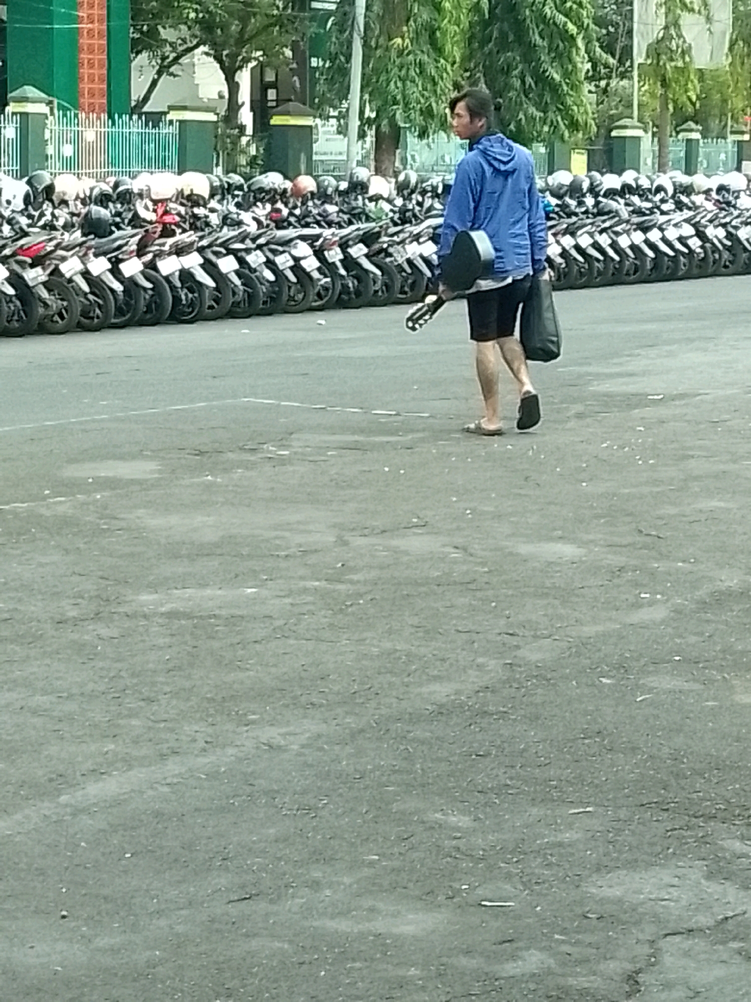 The image depicts a young man walking across a paved area, possibly a parking lot or street, in Jember, Indonesia. He is wearing a blue hooded jacket, black shorts, and flip-flops. He is carrying a dark-colored bag in his right hand and a guitar case slung over his left shoulder.
The background is filled with rows of parked motorcycles, suggesting a location where many people use motorcycles for transportation, such as a public venue, office building, or transportation hub. The motorcycles are parked closely together, forming a dense line. Beyond the motorcycles, there are some trees and structures, including a green building with white railings and pillars in the far left of the frame, which could be part of a university campus, government building, or recreational facility. The ground is asphalt with some patches of wear and tear, and scattered debris. The lighting suggests it is daytime, possibly overcast.