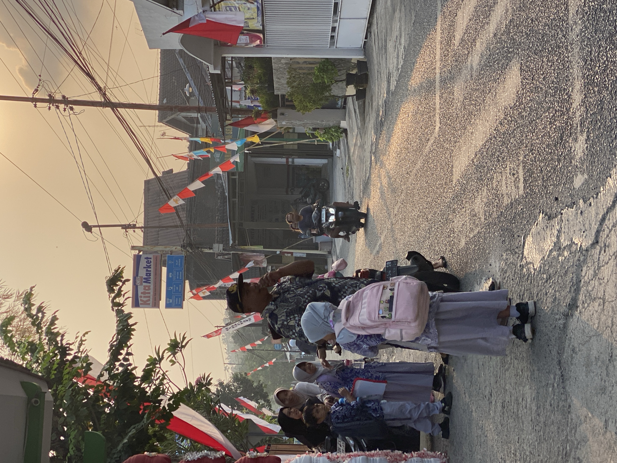 In Kasihan, Indonesia, a street scene unfolds at dusk, bathed in the warm glow of the setting sun. The atmosphere is festive, with numerous red and white Indonesian flags adorning the street. A group of people, including children in school uniforms and adults, are gathered on the side of the road. One child, a girl wearing a light purple headscarf and dress, stands prominently in the foreground, carrying a pink backpack. A man in a dark patterned shirt and a cap, possibly a local official or security personnel, stands nearby, holding a microphone. Motorbikes are present, with one carrying two passengers moving down the street. Shops and houses line the road, with signage indicating a "Kita Market." The street itself is paved, with evidence of wear and tear, and sunlight reflects off the wet surface, creating a shimmering effect. The overall impression is one of a community preparing for or celebrating an event, possibly a national holiday given the prevalent flags.