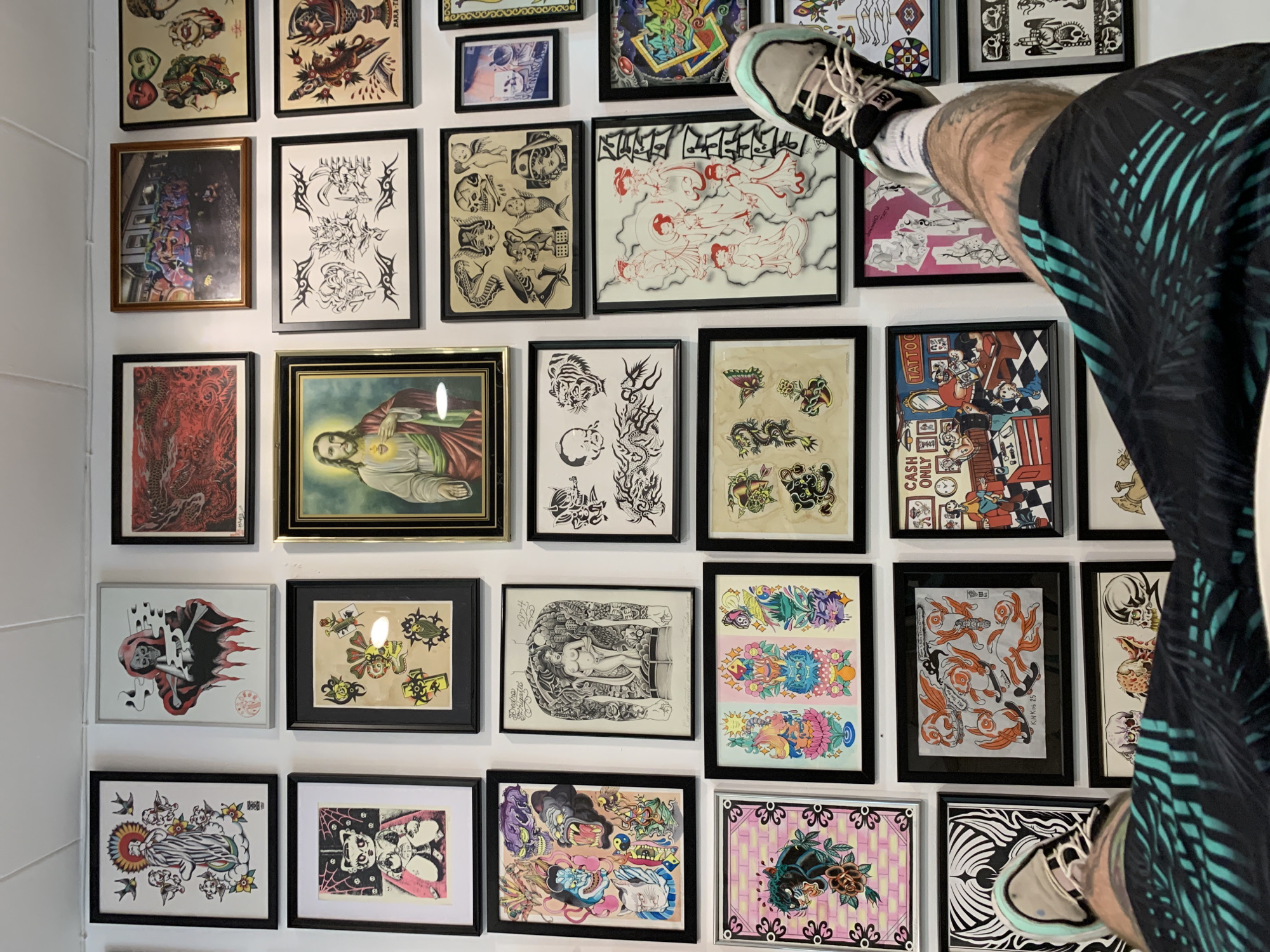 The image displays a densely covered white wall featuring a large collection of framed artworks, primarily tattoo flash sheets, in what appears to be an indoor setting in Barcelona, Spain. On the right side of the frame, the lower torso and legs of a person are prominently visible, clad in dark shorts with a light blue/green tropical pattern, white socks, and light grey sneakers with white laces and mint green soles. One foot is elevated and rests against the wall, suggesting the person is standing on a surface or leaning.
The wall is a mosaic of art, with frames of various sizes and materials, including black, gold, and silver, arranged closely together. The artworks encompass a broad spectrum of tattoo styles, ranging from traditional American and Japanese motifs to intricate blackwork, tribal designs, and vibrant, colorful illustrations. Specific pieces include: a gold-framed photograph of graffiti art; a religious painting of Jesus with the sacred heart; black and white designs featuring skulls, mermaids, and tribal patterns; and dynamic, colorful compositions with dragons, panthers, koi fish, mythical creatures, and floral elements. One striking piece portrays a reaper figure interacting with playing cards, while another depicts a bustling tattoo shop scene.
Visible text within the artworks includes "TATTOO" and "CASH ONLY" on the tattoo shop scene. A large piece with a white background features stylized red text that appears to read "SUNDAY CRITICAL." The text "KINKOS '23" is visible on a black and white artwork featuring koi fish. Several pieces also contain small, sometimes illegible, signatures, dates, or Japanese characters. The overall impression is an eclectic and vibrant display of tattoo artistry.
