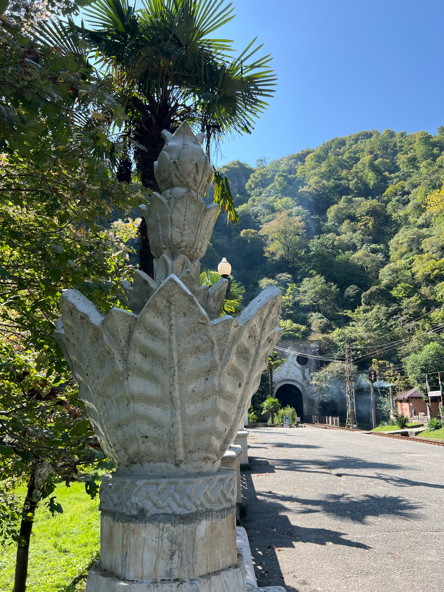 A sunlit, asphalt road curves towards a tunnel entrance in a mountain landscape. In the foreground, a portion of a decorative, pale concrete pillar with lotus-like petal detailing is visible. To the left, lush green foliage and a palm tree are present. The road is shaded by palm fronds. The tunnel, a weathered stone structure with a circular window above its arched entrance, is situated at the base of a densely forested mountain. Power lines and a traffic light are also visible near the tunnel. A small, rustic building with a brown tiled roof stands on the right side of the road. The scene is bathed in sunlight, casting distinct shadows on the road.