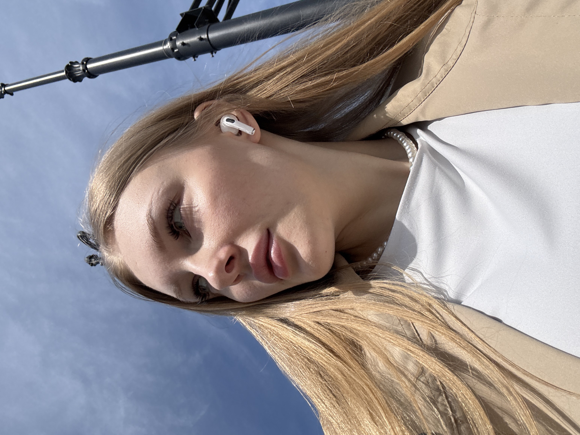 The image presents a close-up, tilted portrait of a young woman outdoors in Perm, Russia. She has long, straight blonde hair and fair skin, illuminated by natural sunlight. A single white wireless earbud is visible in her right ear. She wears a white top, underneath which a pearl necklace is visible. Over her top, she wears a light beige or tan trench coat or jacket. A dark hair clip is partially visible on the left side of her head, blending with her hair.
The woman's gaze is directed downwards and slightly to her left, with a serene and contemplative expression. Her lips are softly parted. The background is a bright, clear blue sky with minimal, wispy white clouds, indicating sunny weather. A dark grey metal pole or structure, possibly a street light or part of a larger fixture, diagonally traverses the upper part of the frame, contrasting with the sky. The bright illumination suggests it is daytime, likely morning or afternoon. No specific activity beyond posing for the photograph is depicted. There is no visible text in the image.