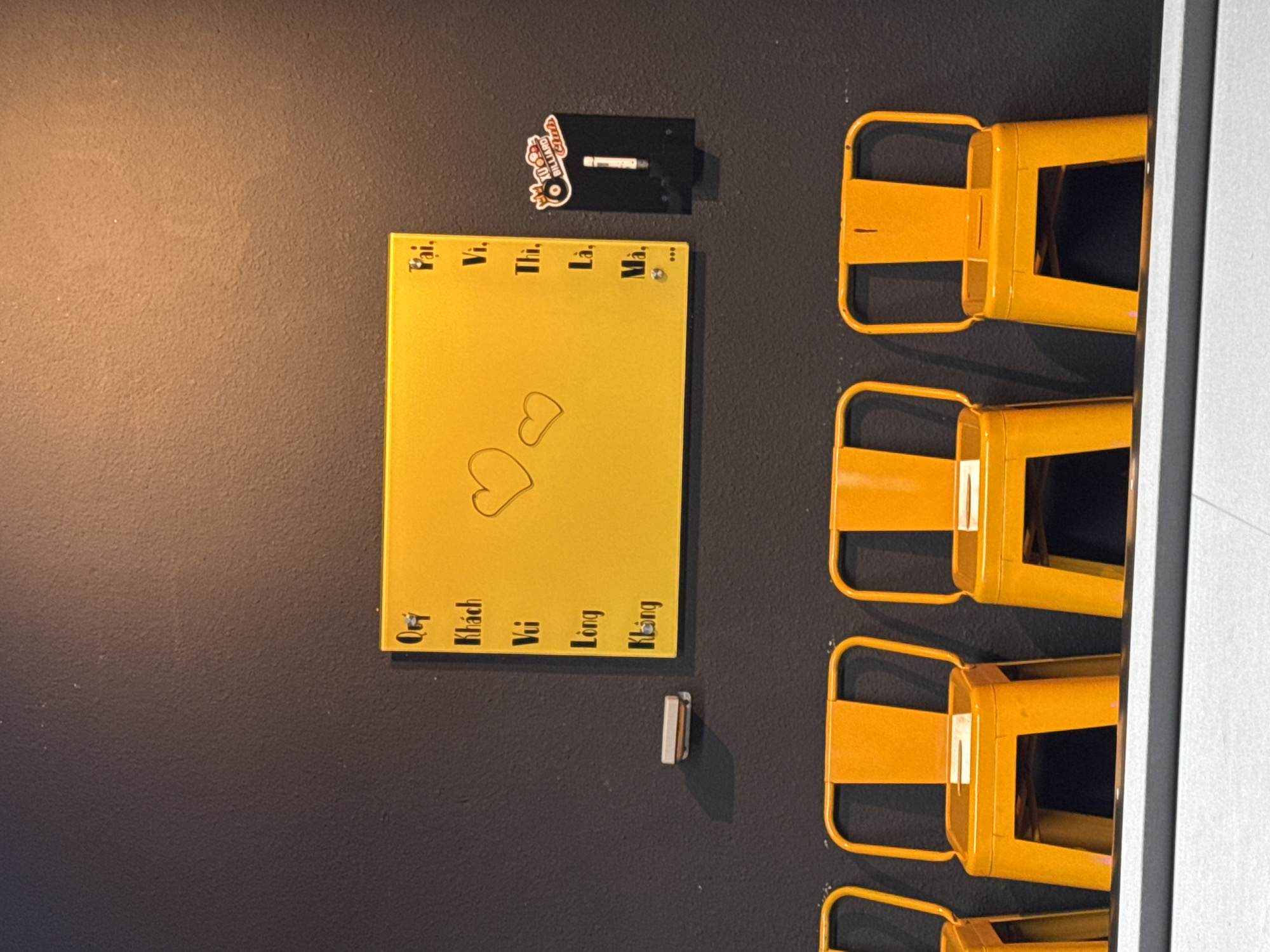 A medium shot depicts a section of a wall with attached objects. The wall appears to be painted a dark, matte gray. On the left side of the image, a bright yellow sign with Vietnamese text is visible. The text includes words like "Khách," "Vui," "Lòng," and "Không," vertically arranged on the left side of the sign. Two heart shapes are drawn on the sign's surface. Above the sign, there's a small black holder with a white object inside. A sticker is affixed to the left of this holder. To the right of the yellow sign and holder, multiple yellow metal chairs are attached to the wall in a vertical row. The chairs appear to be a decorative element. Under the yellow sign is a white object attached to the wall. The overall impression is of an artistic or decorative wall arrangement. The Vietnamese text on the yellow sign, and the fact that the objects are typical of a local shop, suggests that the photograph was taken in a Vietnamese city.