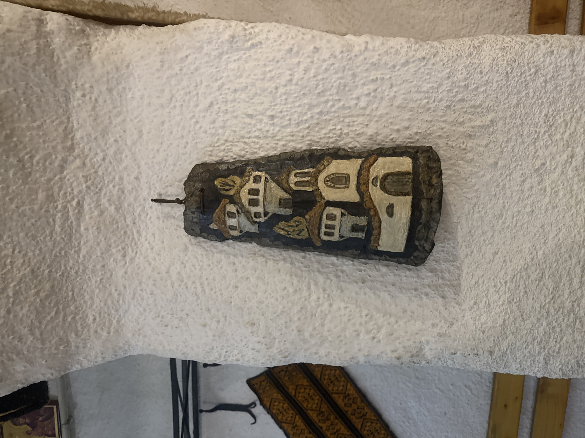 A low-angle, medium shot shows a white, textured ceiling with a piece of art attached. The artwork is a rectangular wooden plaque, painted with images of stylized houses in tan and white, set against a dark background. A small metal hook or pin protrudes from the lower left corner of the plaque. Below the ceiling, the background reveals portions of other objects: a dark-colored painting or piece of furniture, a black metal decorative item, and a patterned fabric. The image appears to be taken indoors, possibly within a building located in Varna, Bulgaria.