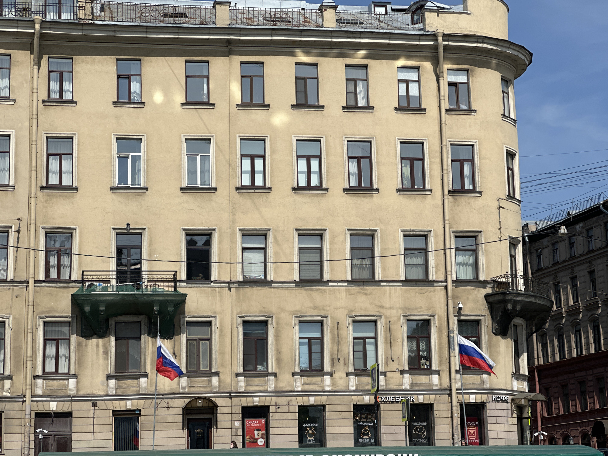 The image shows the facade of a building in Saint Petersburg, Russia. The building has a tan or beige-colored exterior with multiple rows of windows, some with brown frames and others with white. A balcony with green netting hangs from the second floor, along with a Russian flag displayed at both ends. Street-level establishments are visible at the base of the building, with shop signs in Russian, including "ХЛЕБНИК" (Bakery) and "КОФЕ" (Coffee). Some other writing says "СКИДКА Х ДЛЯ СТУДЕНТОВ" and other signage related to a coffee shop that has discounts for students. Other building elements include a rounded corner on the right side, typical of Saint Petersburg architecture. A metal railing is visible on the roof. In the background, parts of other buildings are seen, along with some blue sky.