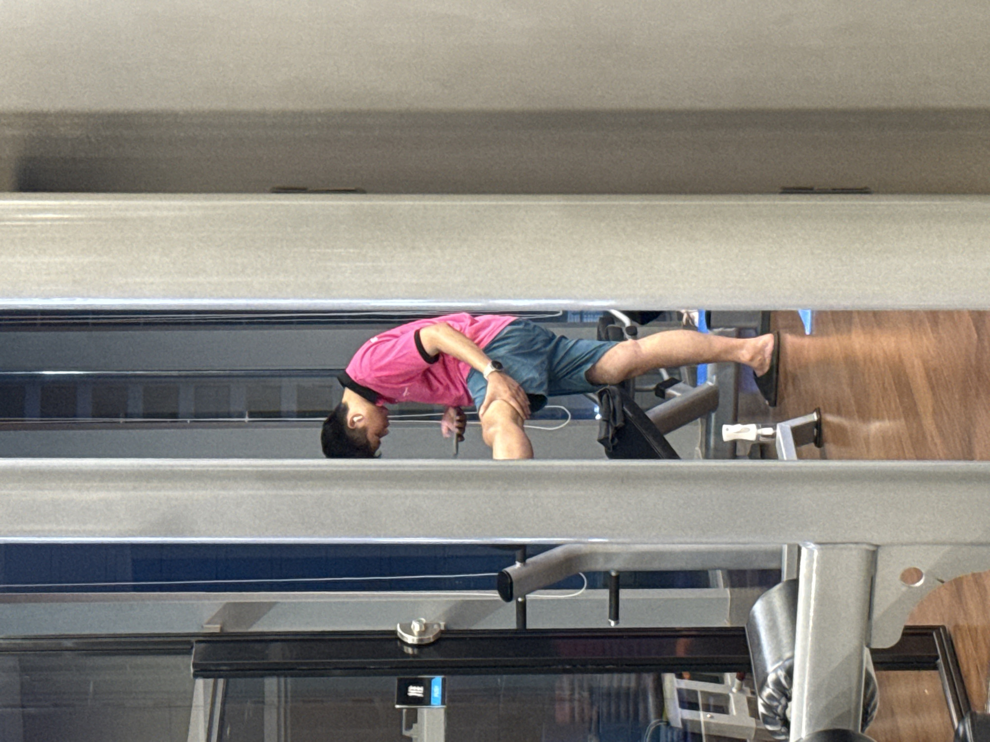 The image shows a person in a gym, seemingly engaged in a stretching or exercise routine. The person is wearing a pink t-shirt and blue shorts. Their body is contorted in a way that suggests a deep stretch, with their head bent down towards their chest and their legs extended. The gym environment is visible, with exercise equipment and wooden flooring. The scene is lit by artificial light, suggesting it could be any time of day. The image appears to be taken from a slightly elevated angle, looking down at the person. There are no clear indicators of the time of day or weather conditions. The overall impression is one of a person focused on their physical activity within a fitness setting.
