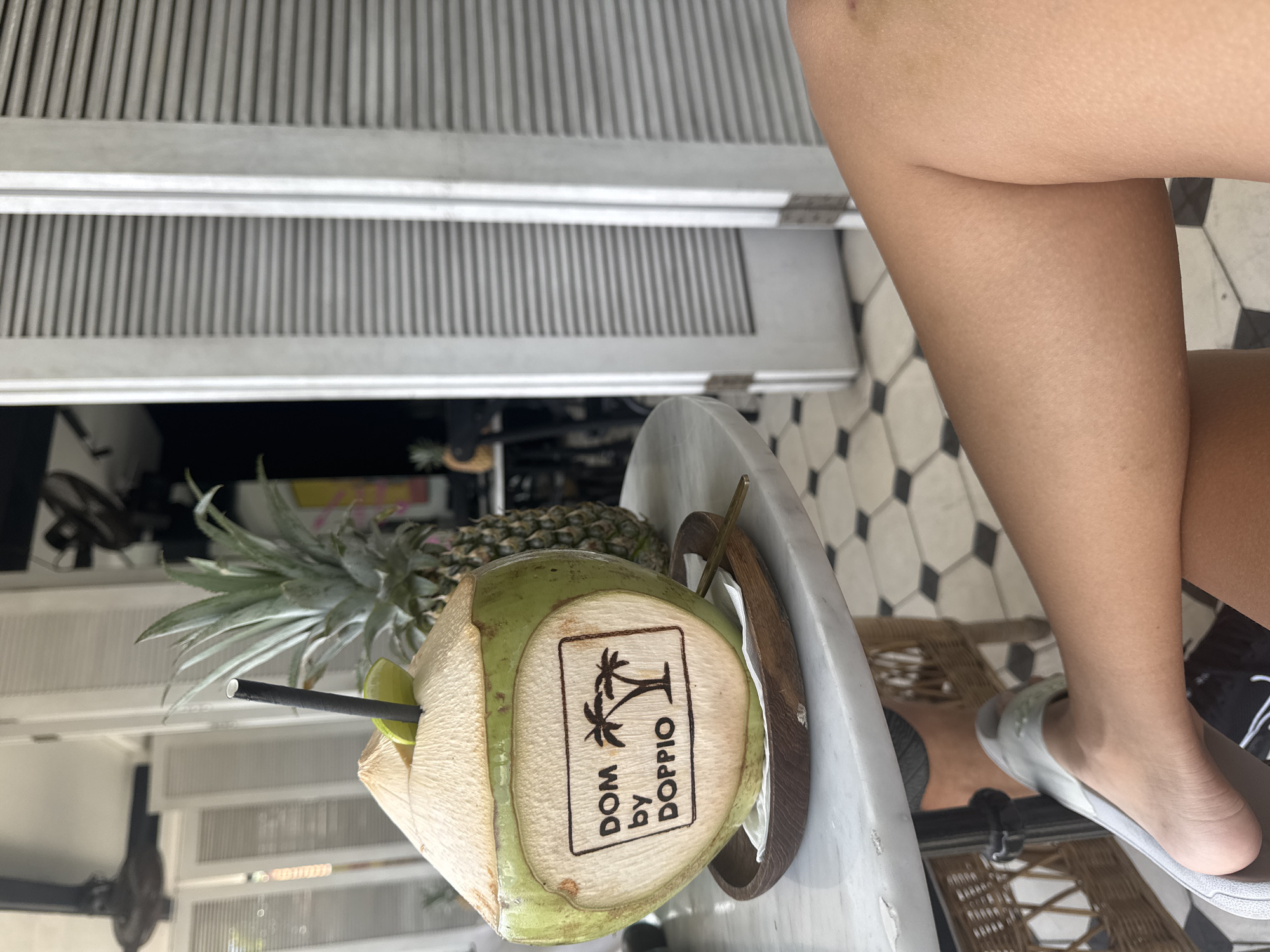 A view from above captures a person seated next to a small, round, marble table in what appears to be a cafe or restaurant. On the table is a fresh coconut drink, decorated with a logo that reads "DOM by DOPPIO", a pineapple resting on the table behind the coconut, and a straw inserted into the coconut. The background includes a partial view of a white-louvered door and a patterned black and white tiled floor. The person is wearing a pair of light grey slip-on sandals. The location may be North Kuta, Indonesia, given the label on the drink.
