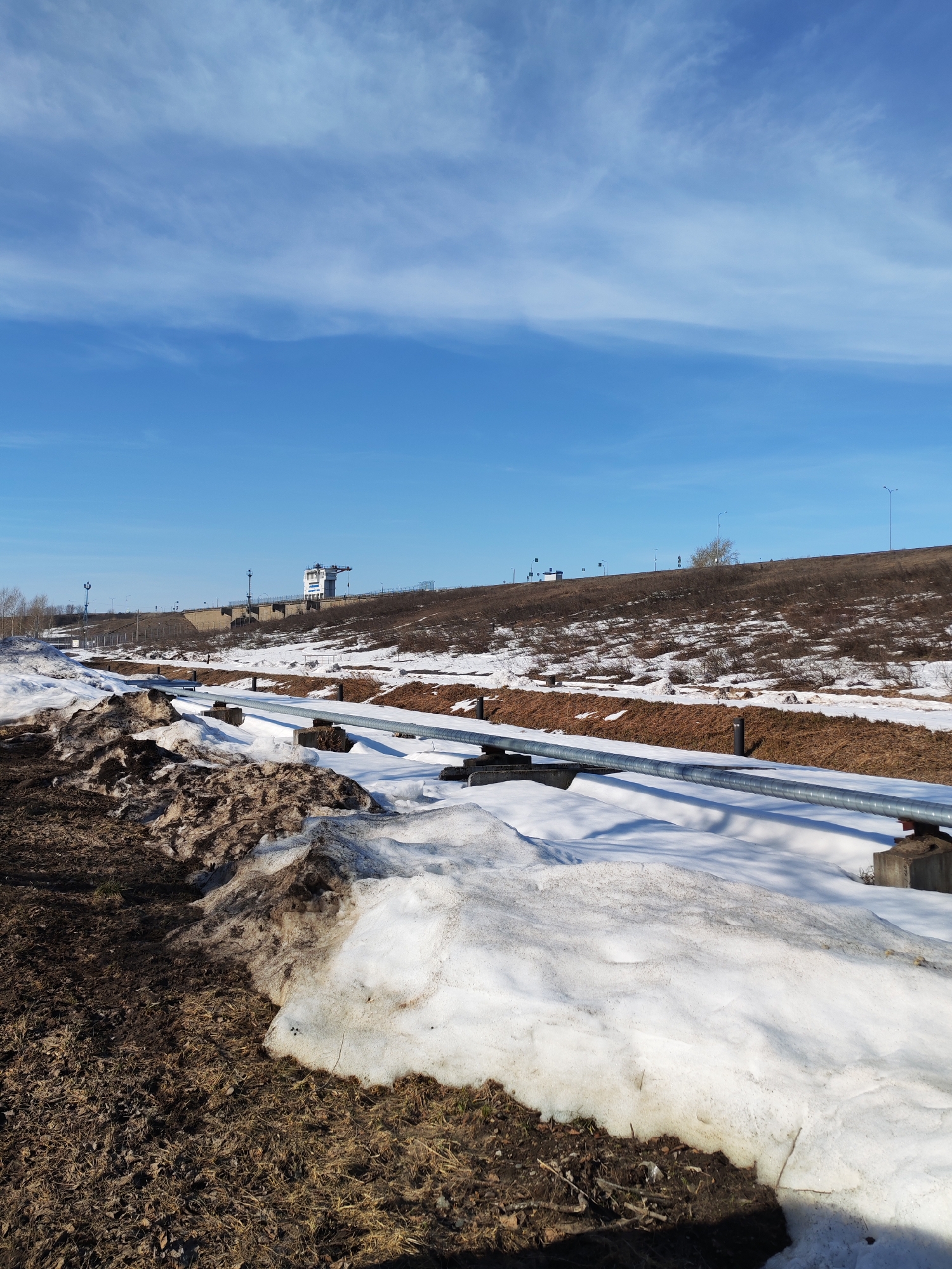 The image depicts a landscape in Chaikovsky, Russia during the transition from winter to spring. The foreground shows thawing ground with patches of snow and exposed brown earth. A gray, cylindrical pipeline runs horizontally across the middle ground, supported by concrete blocks. Behind the pipeline is a sloping embankment covered with dry grass and sporadic patches of snow. On the embankment, there's a structure that appears to be a navigational control tower, along with streetlights and other infrastructure. The sky is blue with scattered clouds. The scene suggests early spring conditions with remnants of winter snow melting away. There are no people visible in the image.