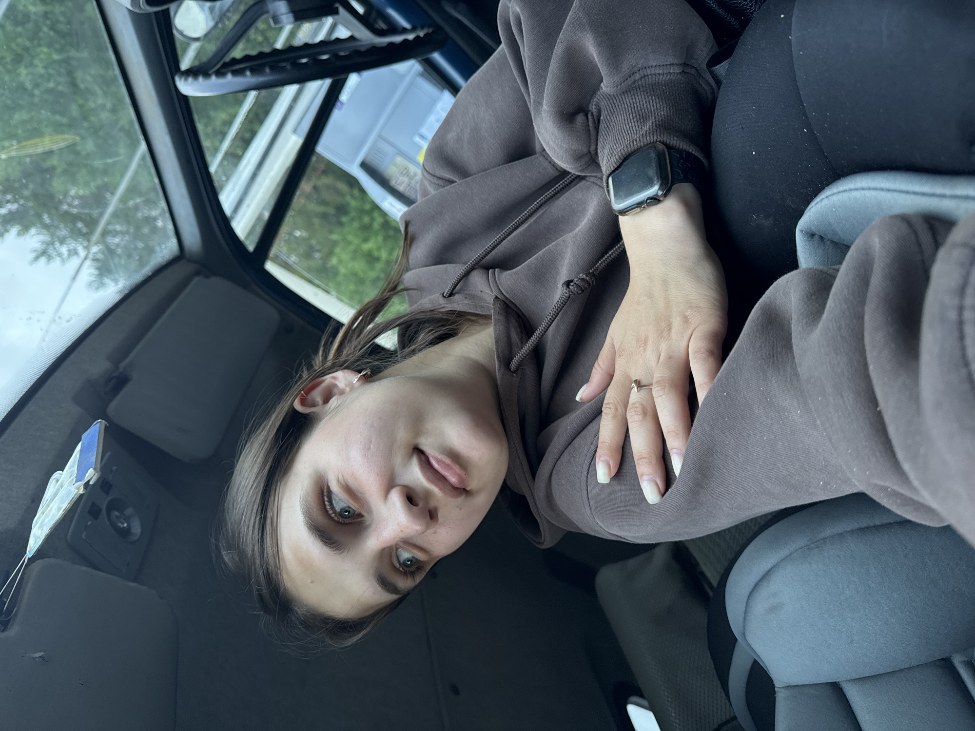 A young woman with light brown hair, wearing a brown hoodie and black leggings, sits in the passenger seat of a vehicle. She is looking to her left, with her left hand resting on her chest and her right hand on her lap, showing a silver ring on her ring finger and an Apple Watch on her wrist. The interior of the car is visible, including a grey car seat, the dashboard, and the windshield. Outside the car, trees and a grey building can be seen. There is no discernible information that indicates this image was taken in Vary, Russia.