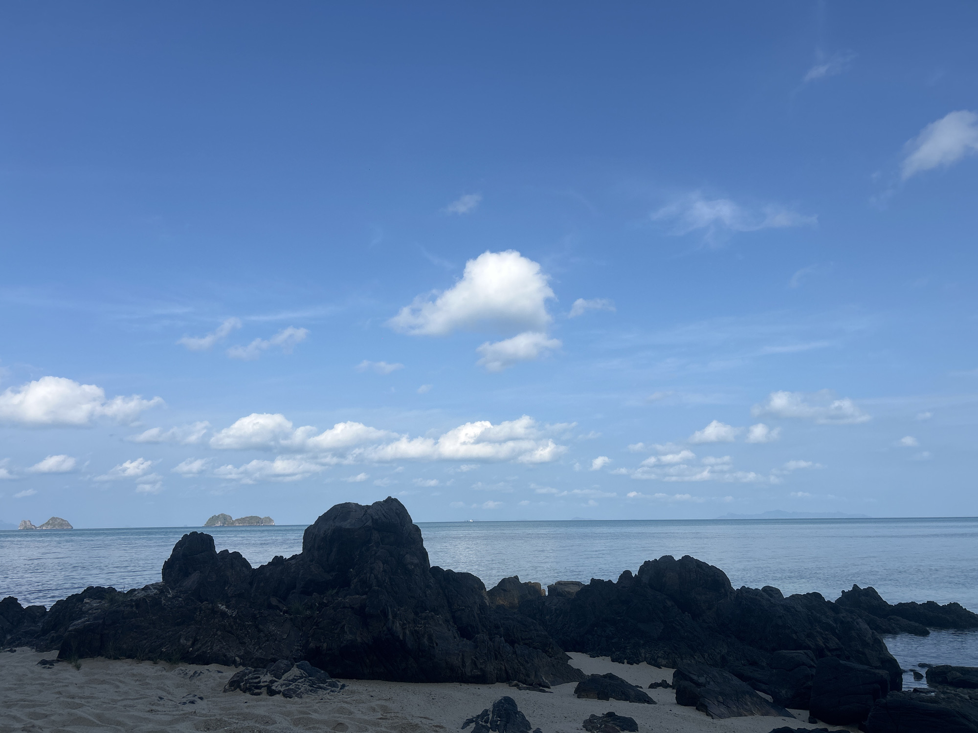 The image presents a serene coastal landscape at Baan Taling Ngam, Thailand. In the foreground, large, dark, rugged rock formations are prominent, contrasting sharply with the light beige sand of the beach. These rocks vary in size and shape, forming an irregular line across the bottom and mid-ground of the composition.
Beyond the rocky shore, a calm, blue-grey ocean extends towards the horizon. The water appears still, with a slight haze or gentle ripple visible. In the mid-distance, several small, dark islands or landmasses emerge from the sea, adding depth to the scene and indicating a typical tropical archipelago setting.
The upper half of the image is dominated by a vast, clear blue sky. Scattered white cumulus clouds are dispersed across the sky, with one notably fluffy cloud positioned centrally in the upper frame. The bright illumination suggests it is daytime, likely late morning or early afternoon, with clear weather conditions.
There are no people or specific activities depicted, emphasizing the natural and untouched quality of the environment. The overall mood conveyed is one of tranquility and natural beauty. No visible text is present in the image.
