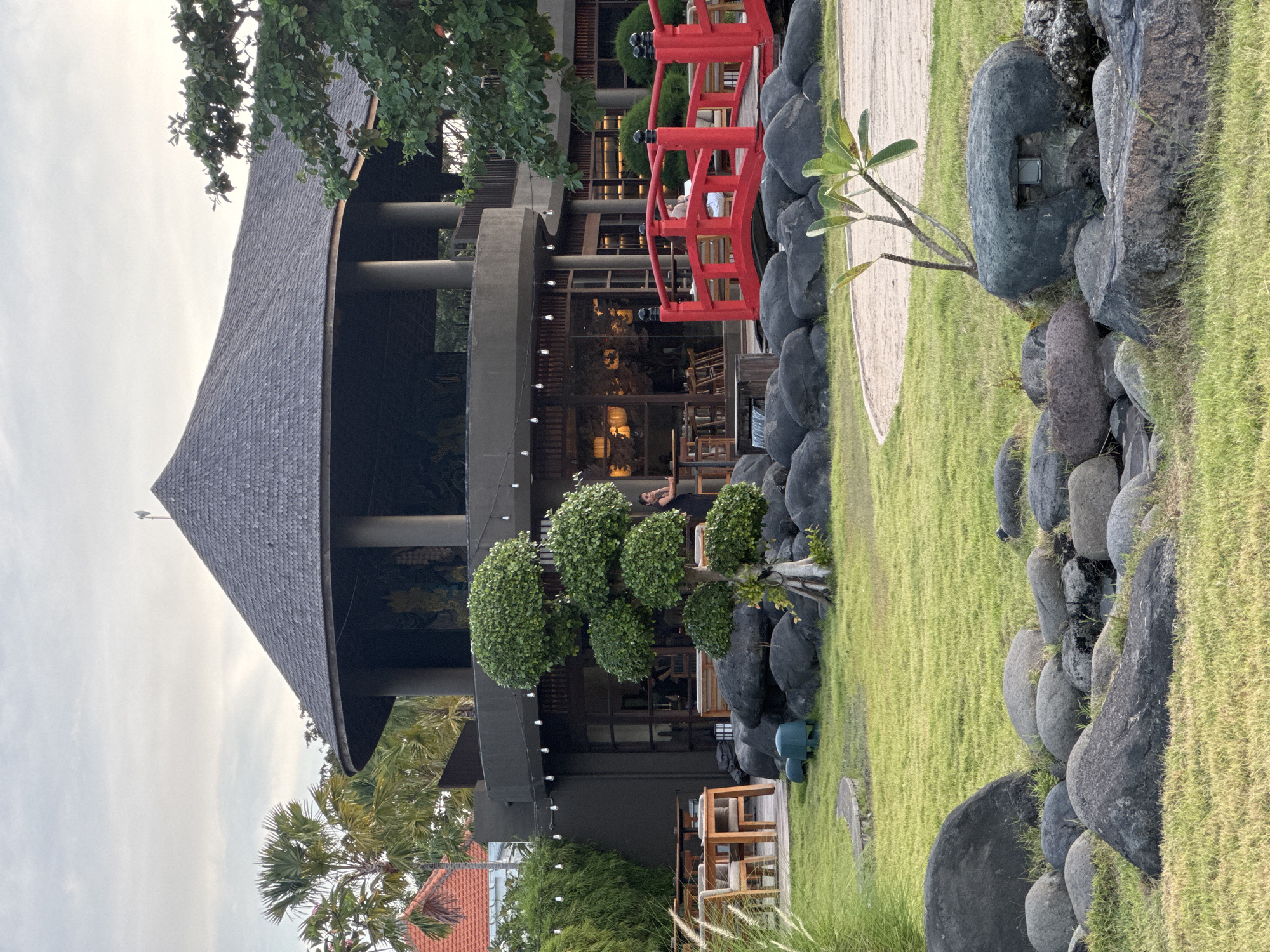 Here is a detailed description of the image:
The image captures an outdoor scene, possibly a restaurant or venue, in a city identified as "Unknown, Unknown." The primary structure is a multi-story building with a distinctive conical roof, suggesting an Asian architectural influence. The building is painted in dark gray and is adorned with string lights.
A red, arched bridge stands out against the dark background of the building. To the right of the bridge is a rock garden arrangement with dark, rounded stones and patches of green grass. A small tree with sparse foliage is planted near the stones.
In the foreground, there is a well-manicured lawn and another ornamental tree shaped like a series of spheres. Further back, there are tables and chairs, indicating an outdoor seating area. Palm trees and other greenery are visible, adding to the tropical atmosphere. The sky above is overcast, creating soft, diffused lighting. A woman is seen standing on the second floor of the building.