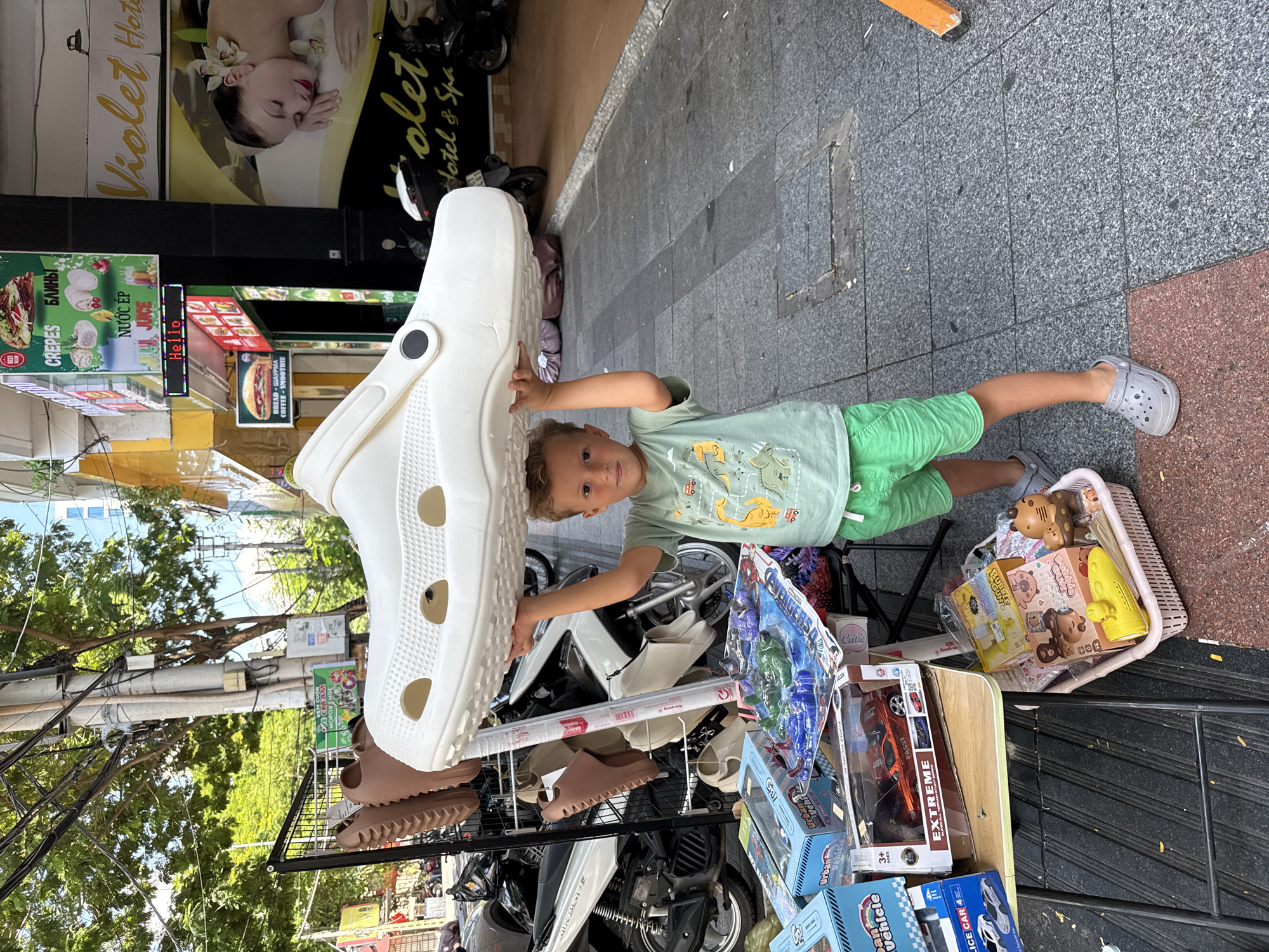 A young boy, approximately 7 years old, stands on a sidewalk in Nha Trang Ward, Vietnam, holding a very large white clog shoe above his head. He is wearing a light green t-shirt with cartoon dinosaurs and bright green shorts. He is also wearing gray clogs. The sidewalk is paved with gray and red tiles. To the left of the boy is a stall with various items for sale, including toy cars in boxes, action figures, and other small toys in a pink basket. Behind the stall, a rack displays several brown clogs. In the background, there are buildings with signs, including "Violet Hotel & Spa." Motorcycles are parked in the background, and trees with green foliage line the street. Overhead, there are many electrical wires and poles. The lighting suggests it is daytime, with sunlight casting shadows.