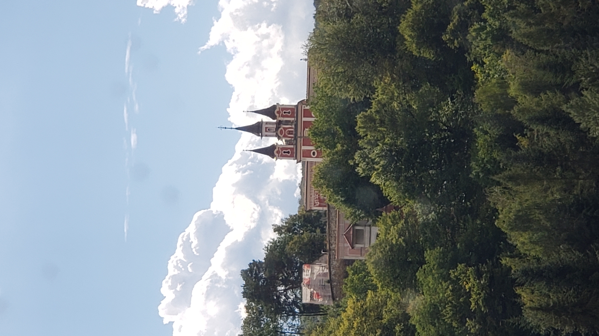 An eye-level, medium shot captures a red and white church building situated on a hill, partially obscured by lush green trees. The church features multiple spires, with the central one being the tallest, and ornate architectural details. The sky above is a bright blue with scattered white clouds. In the foreground, a dense canopy of various trees dominates the lower portion of the image. To the left of the church, a construction site is visible, with scaffolding and materials suggesting ongoing renovation or building work. The scene is set in the city of Prešov, Slovakia. There are no people or animals visible in the image.