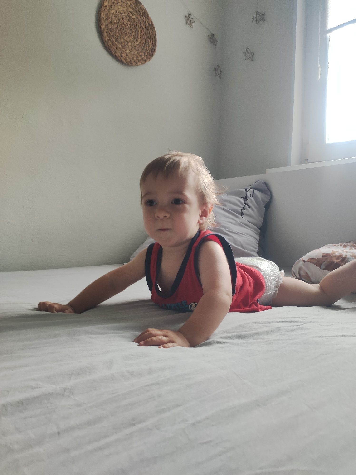 A toddler lies prone on a bed covered with a light gray sheet. He wears a red sleeveless shirt and a diaper. His arms are extended in front of him, and his legs stretch out behind. Behind him are pillows and a white wall, with a window to the right. On the wall are a circular woven decoration and a string of star-shaped decorations. The scene appears to be indoors, likely a home. The location of the scene is not possible to determine without additional information.