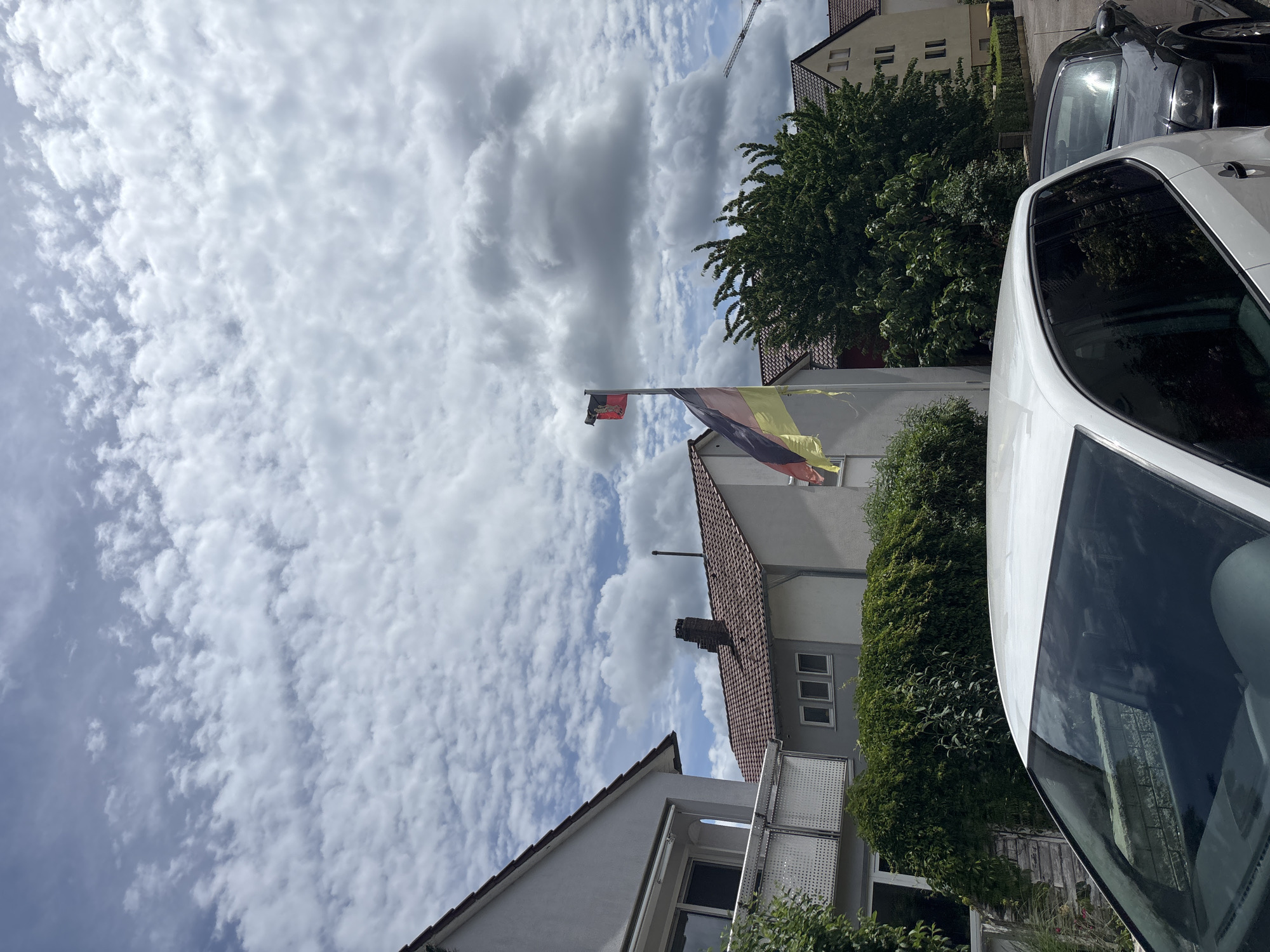 In an outdoor urban setting, two cars are parked on a street. The car in the foreground is white, and the car in the background is black. A German flag is flying on a flagpole. In the background, several houses are visible, with trees and a cloudy sky overhead.
