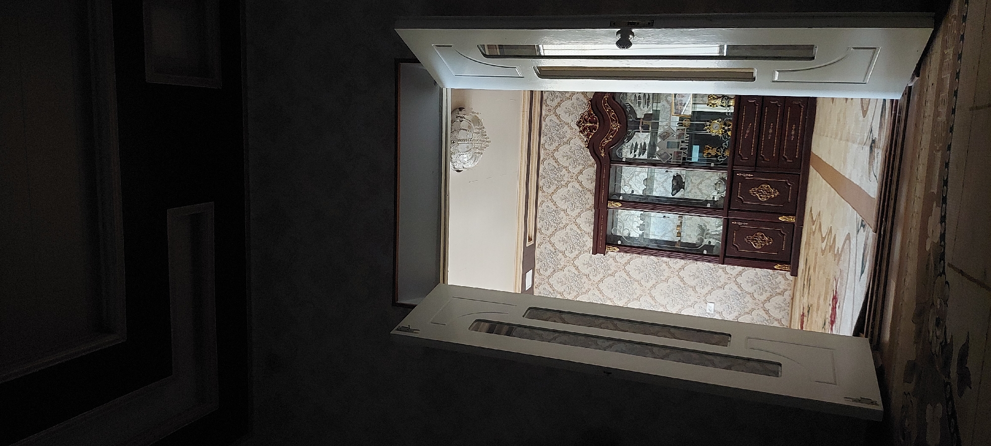 The image depicts an interior scene, likely a living room or dining room, viewed from a darkened hallway through an open doorway. Two white doors, partially ajar, frame the view into the illuminated room. The left door is angled inward, revealing a decorative inlay. The right door is open wider, showing a doorknob and a paneled section.
In the background of the room, a large, ornate, dark wooden display cabinet with glass doors is prominently featured. The cabinet is filled with various decorative items, including glassware, figurines, and what appear to be clocks. The cabinet sits against a wall with patterned wallpaper featuring a floral or damask design in neutral tones. Above the wallpaper, a dark border runs along the ceiling. A crystal chandelier hangs from the ceiling in the center of the room.
The floor is covered by a patterned carpet with light and dark beige tones and hints of red. The lighting in the room is bright, contrasting with the dark hallway in the foreground, which has a dark, textured ceiling with recessed geometric elements. The overall impression is of a well-furnished and decorated interior space. The location is identified as Shamirza, Uzbekistan.