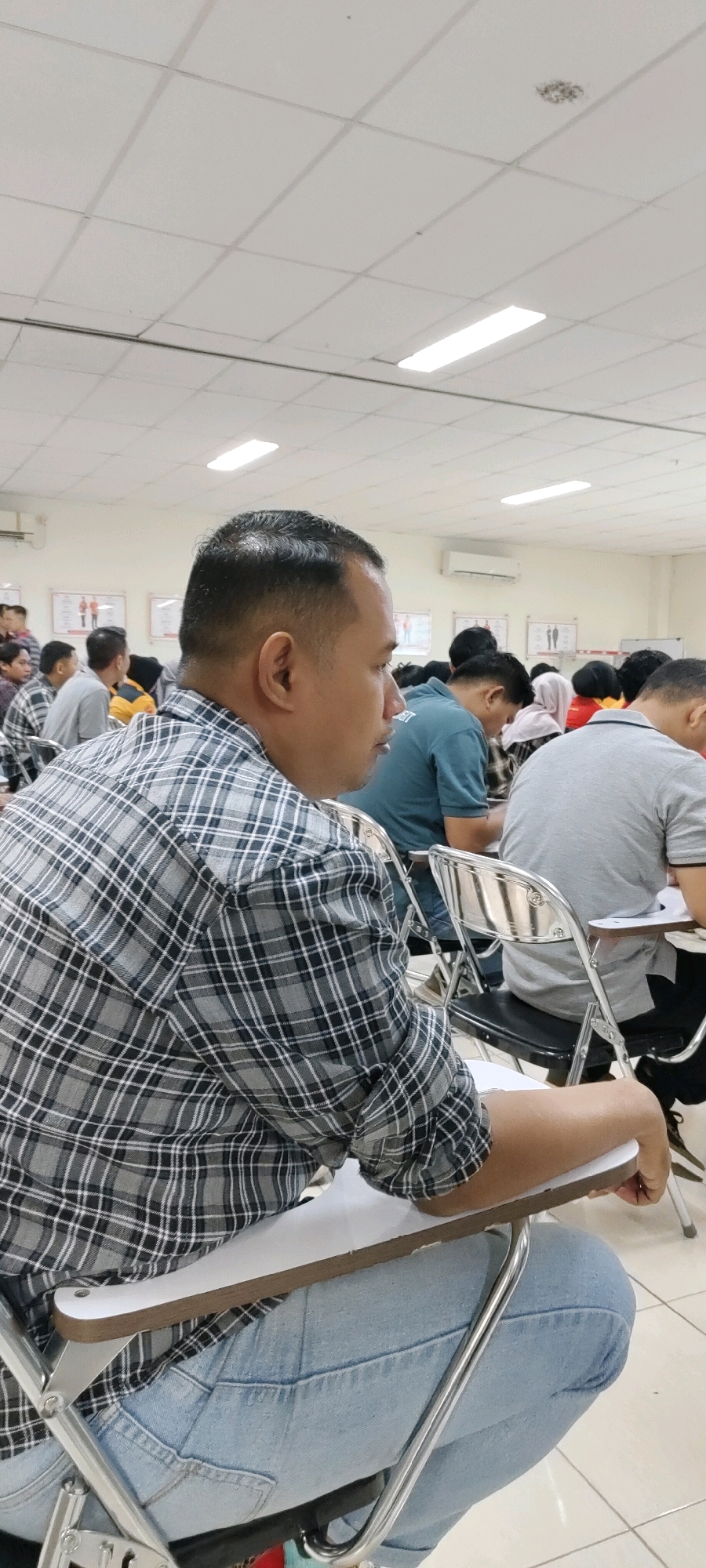 The image depicts an indoor classroom setting with several individuals seated at desks, facing forward, suggesting a lecture or examination. In the foreground, a man with short hair is seen from the side, wearing a grey and black plaid shirt and blue jeans. He is seated in a folding chair with a writing tablet attached. Behind him, other students are visible, mostly with their backs to the camera. They are also seated in similar chairs and appear to be attentive. The room has a tiled ceiling with fluorescent lighting panels and white walls. Posters depicting human figures are displayed on the back wall, possibly related to the subject being taught. An air conditioning unit is mounted high on the wall. The overall environment suggests a formal educational institution. The image does not provide any specific landmarks or clues to pinpoint the exact location within the city of Nusa Indah, Indonesia.