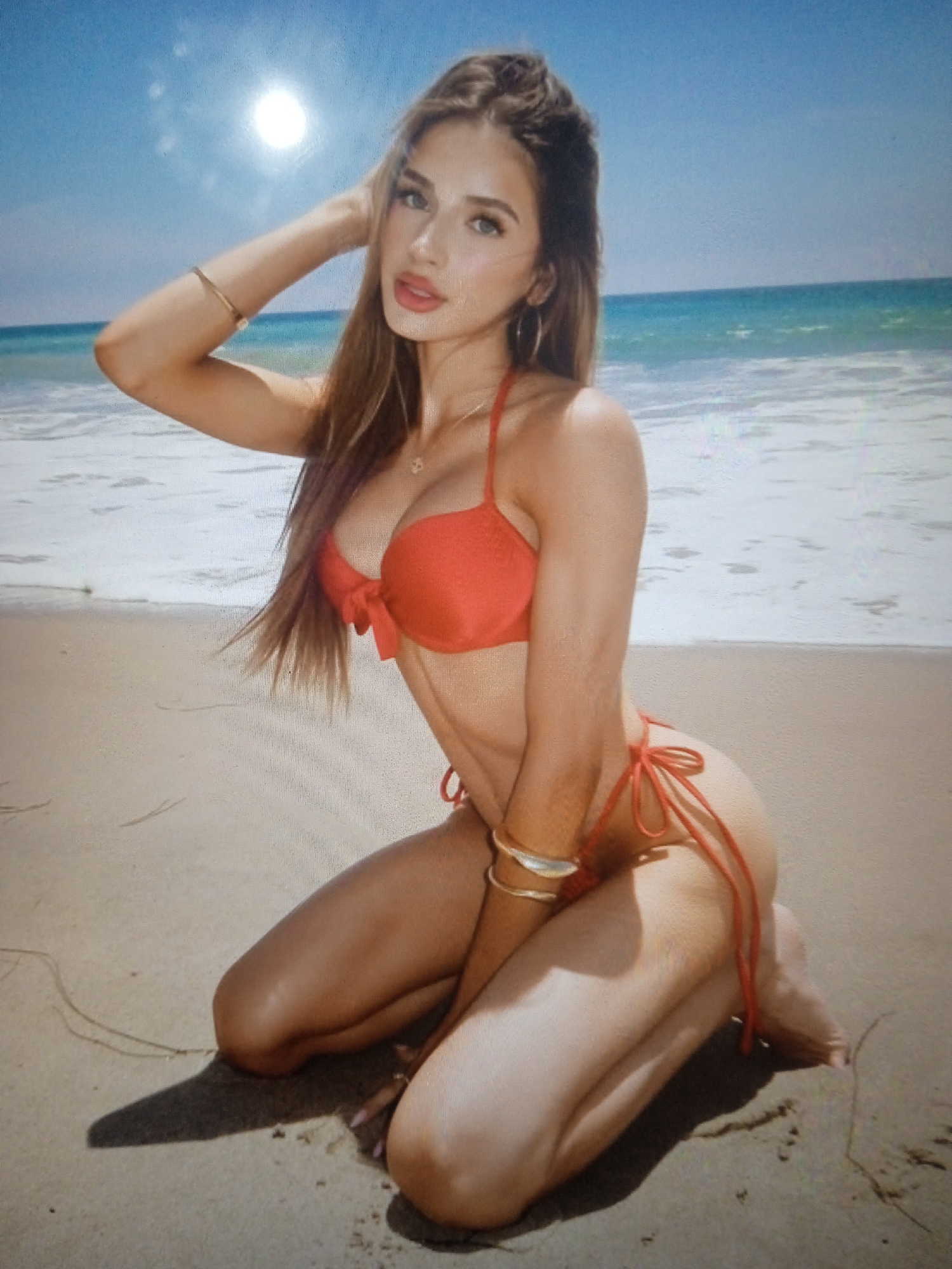 The image shows a woman in a red bikini kneeling on a sandy beach with the ocean and sky in the background. The woman is positioned in the foreground, facing the viewer. She has long, brown hair and is wearing bracelets on her left arm. Her right arm is resting on her head. The beach appears to be a natural setting, possibly a coastline. There are no visible elements in the image that suggest a connection to the city of Chernivtsi, Ukraine.