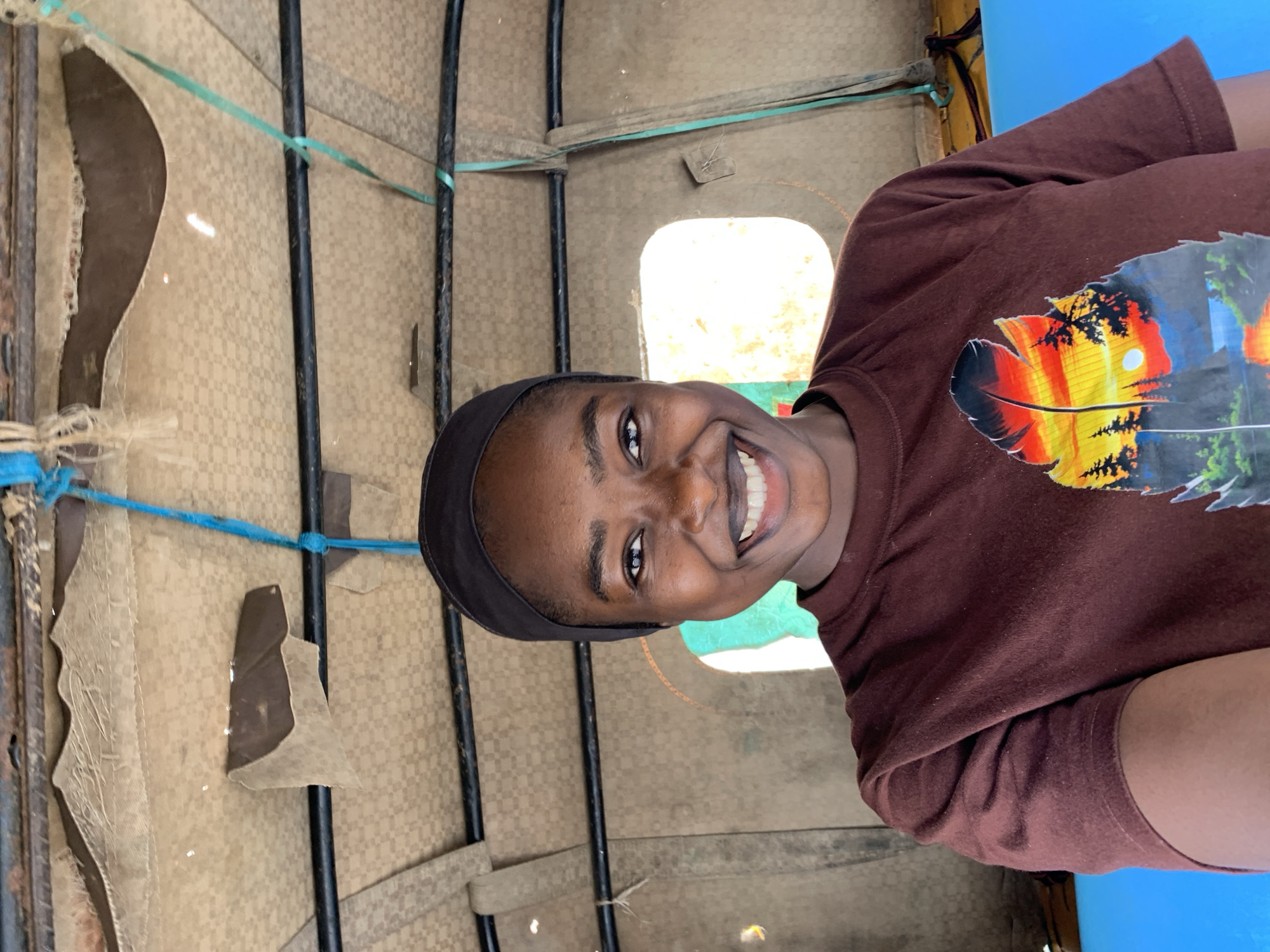 A young woman is in what appears to be a "Keke Napep" or auto rickshaw in Rafin Par, Nigeria. The vehicle's interior has a worn and patched beige covering. She is smiling broadly at the camera and wearing a dark headband and a brown t-shirt with a colorful feather design on it. The backdrop is the interior of the vehicle, with glimpses of the outside visible through the windows.