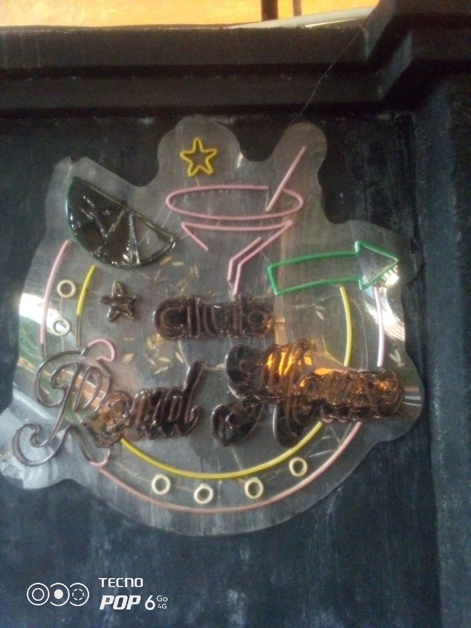 The image shows a neon sign for "Royal Club" in Howayi, Nigeria. The sign is shaped like a rounded rectangle with a star at the top and a slice of lime. The words "Royal Club" are written in a cursive font. The sign is illuminated with pink, yellow, and green neon lights. The sign is mounted on a dark wall. A Tecno Pop 6 4G logo is visible in the bottom left corner of the image.