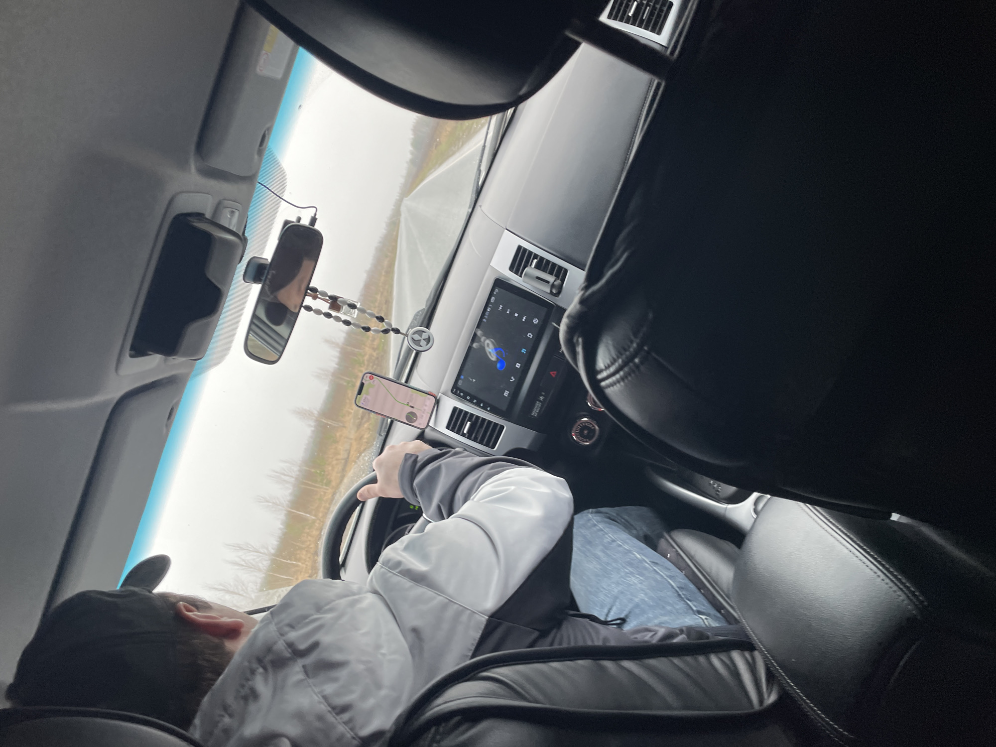 The image shows the interior of a vehicle, specifically the driver's seat and dashboard. A person wearing a black cap and a gray and white jacket is seated in the driver's seat with their hands on the steering wheel. A smartphone is mounted on the dashboard, displaying a navigation application. The car's infotainment system screen is visible between the air vents. A rearview mirror with a beaded ornament hanging from it is present. The background visible through the windshield suggests an overcast day in a rural area, possibly a road with trees on either side. Since Sidorovy Gory, Russia, is primarily a rural area, the scene likely depicts a local road.