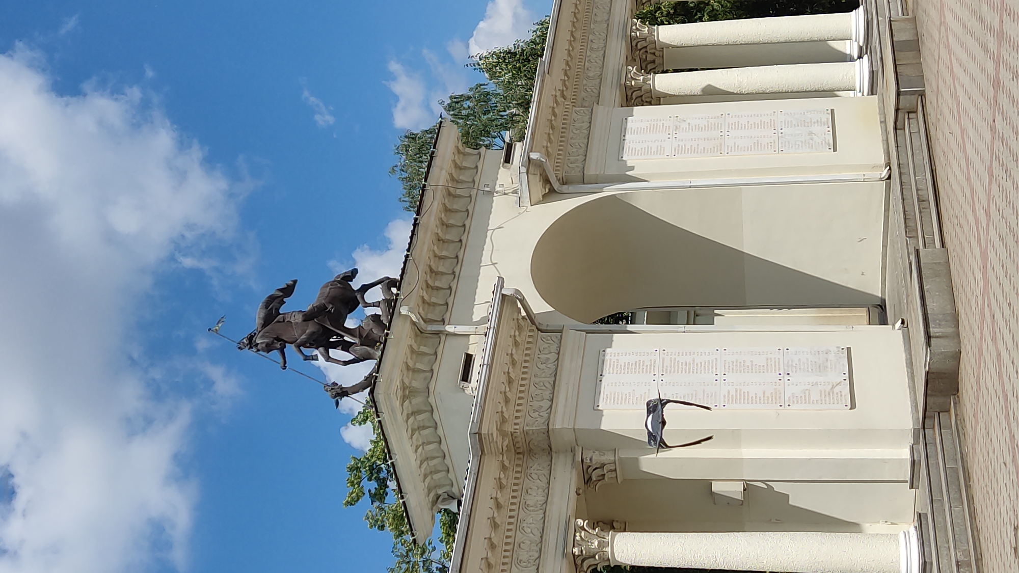 The image captures a monument in Krasnodar, Russia, depicting a bronze sculpture of St. George slaying a dragon, atop an archway. The archway is constructed of cream-colored stone with classical architectural elements, including Corinthian columns and decorative friezes. Two memorial plaques are affixed to the sides of the archway, listing names. A pair of dark-colored spectacles rests on one of the plaques. The sky is blue with scattered white clouds, and lush green trees are visible behind the monument. The ground in front is paved with patterned brickwork, and there are stone steps leading up to the archway. The overall scene is serene and suggests a public space.