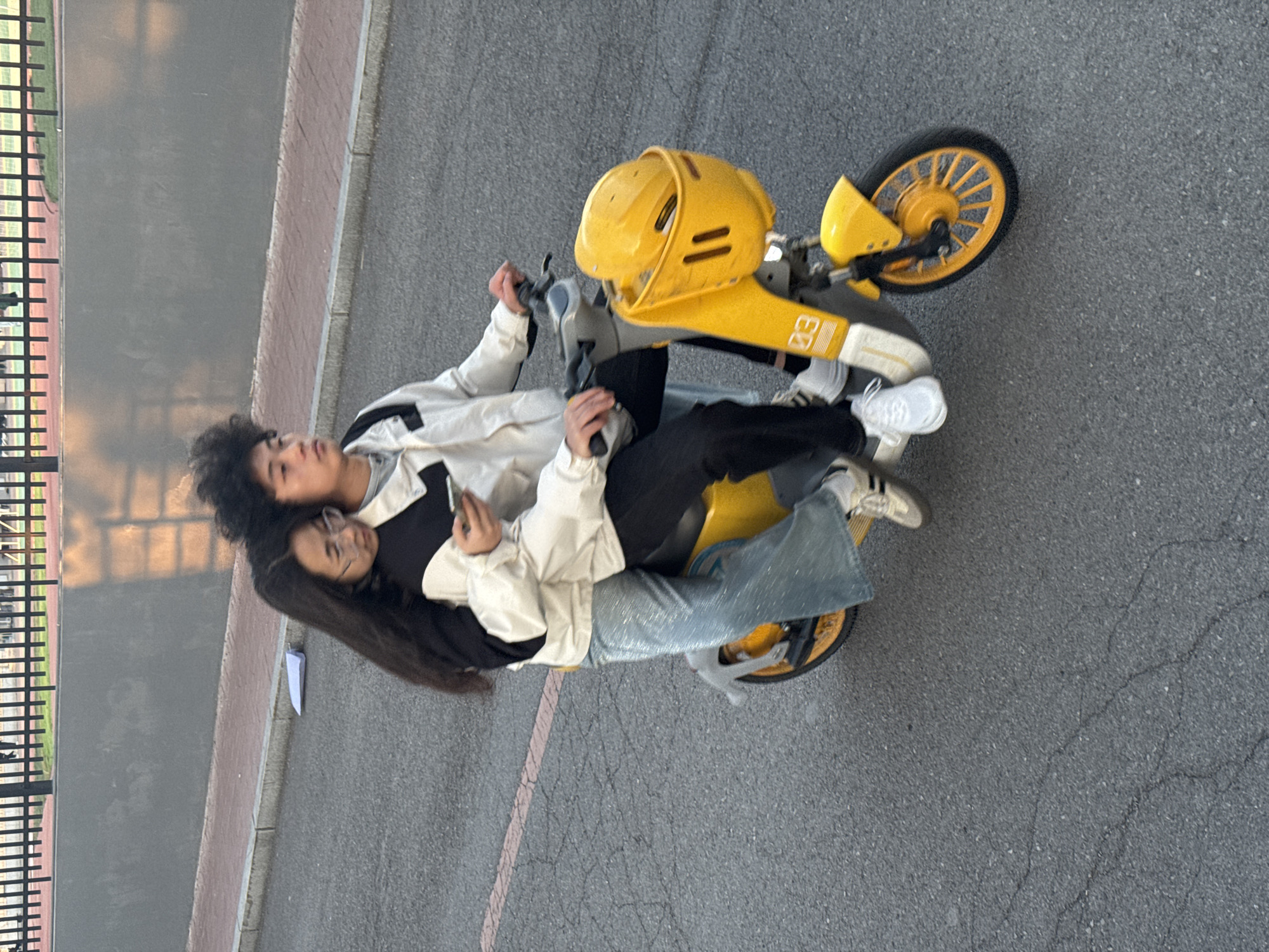 A woman and a man are sitting on a yellow scooter on an asphalt surface. The man is in the front and operating the scooter. The woman is sitting behind him, holding a phone, and resting her head against his back. A metal fence is visible on the left of the image. The location is Zhongtun, China.