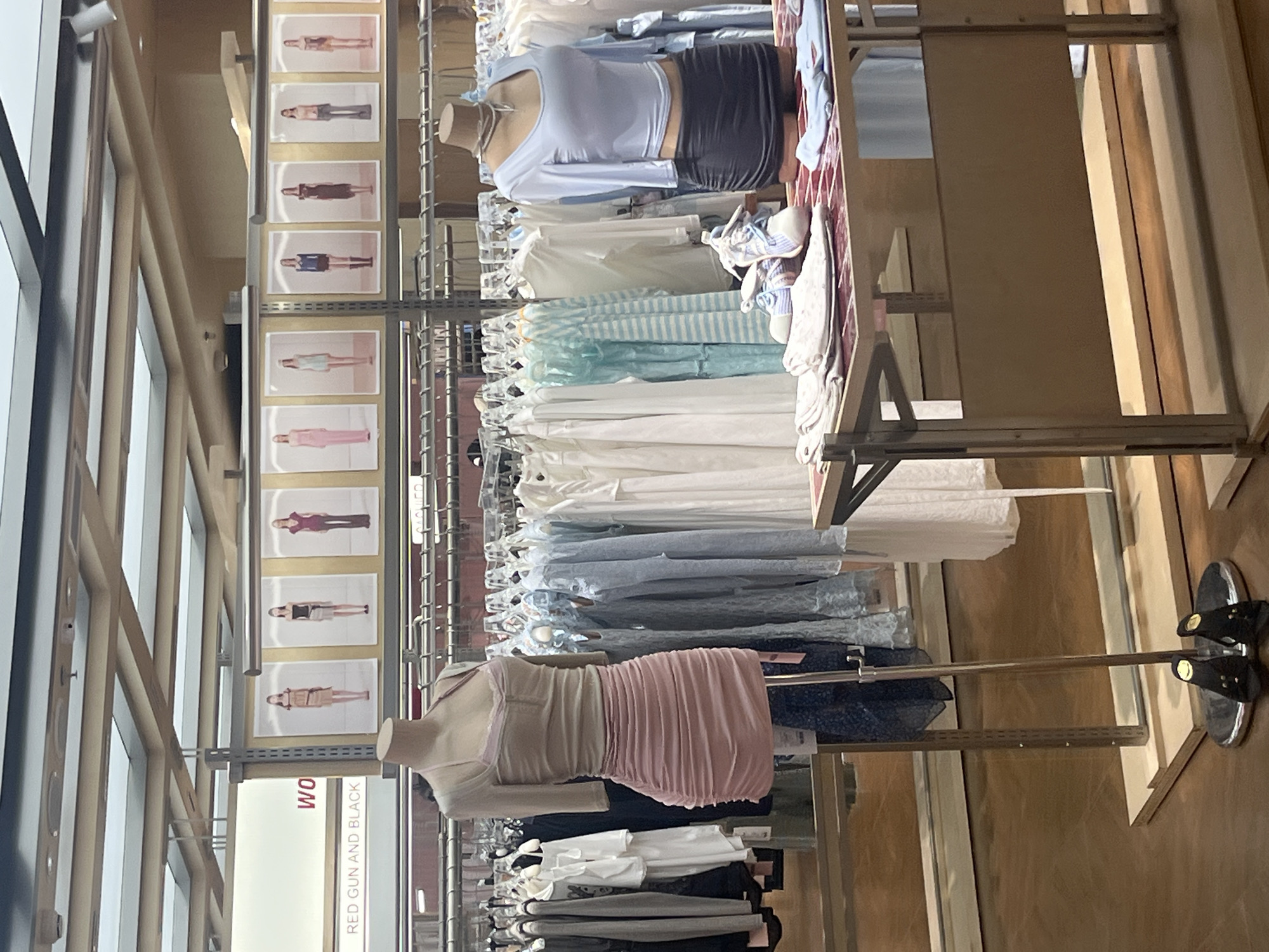 The image depicts an indoor clothing store, likely in Yuzhong District, China, based on the provided context. The scene is characterized by clothing racks filled with garments, mannequins displaying outfits, and display tables.
In the foreground, a mannequin is dressed in a light pink ruched skirt and a light beige halter-neck top. To the right, another mannequin wears a light blue long-sleeved crop top and a dark grey ruched skirt, accessorized with a delicate necklace. In front of this mannequin, a display table holds folded clothing items, a pair of white and blue athletic shoes, and a red-tiled surface. Another display table is visible further back, also with folded clothing.
Behind the mannequins, clothing racks are densely packed with various garments, predominantly in light colors such as white, light blue, and grey. Posters featuring female models in different outfits are displayed on the wall above the clothing racks. A sign with "WO" is partially visible on the left, and below it, "RED GUN AND BLACK" is displayed.
The flooring is a reflective, polished wood-like material. The ceiling features a grid of exposed beams and what appear to be skylights or light panels. In the lower left corner, a pair of black, open-toed shoes with smiley face embellishments rests on a circular base. The overall ambiance suggests a retail environment focused on contemporary fashion.