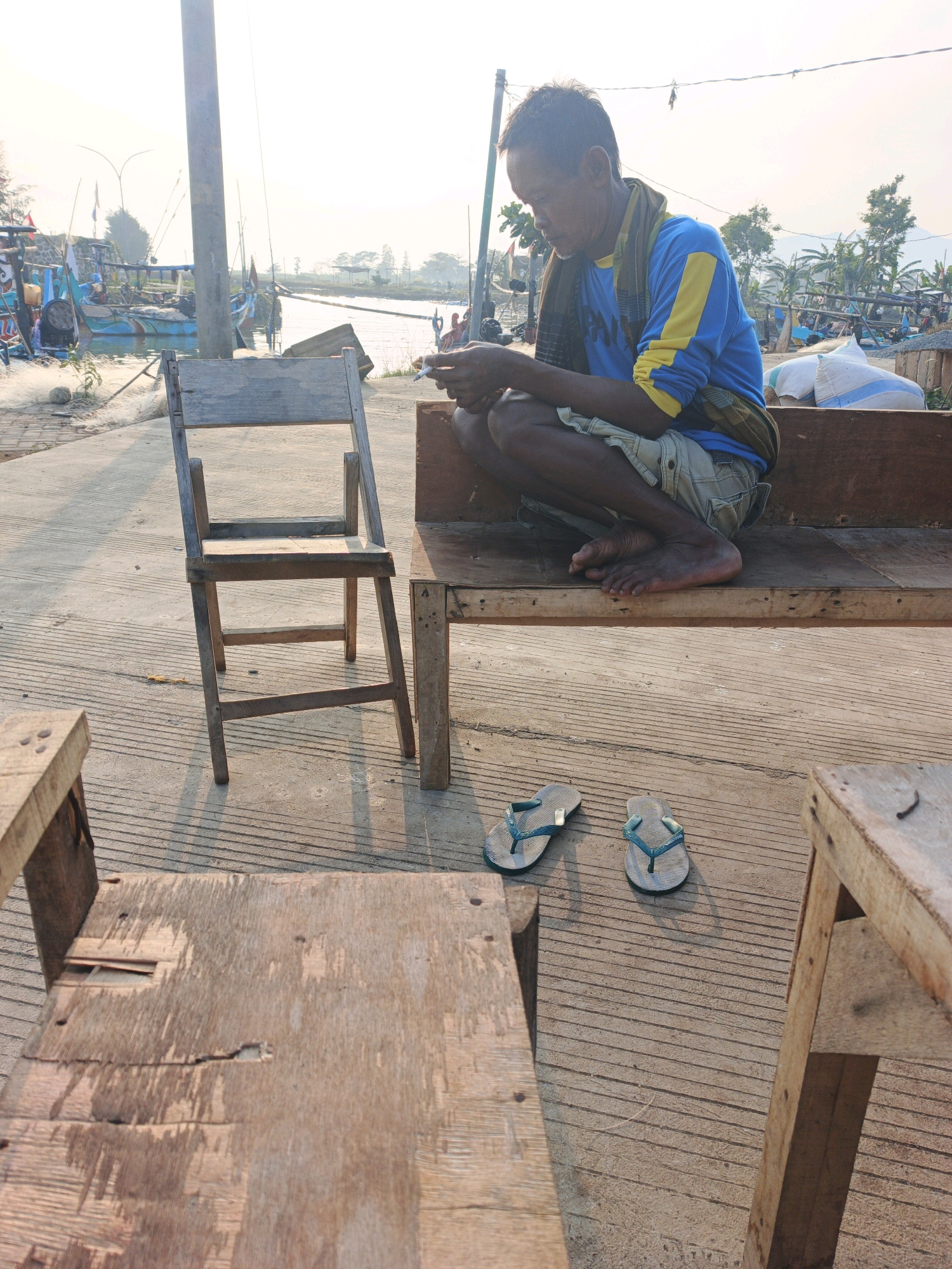 A middle-aged man with dark skin and short, graying hair sits on a wooden bench, looking down at his hands which are holding a smartphone. He wears a blue shirt with yellow accents on the sleeves, beige cargo shorts, and a patterned scarf around his neck. His feet are bare. Two flip-flops, one teal and one a lighter shade of teal, are placed on the concrete ground in front of the bench. To the left of the bench is a small, wooden chair. In the foreground, out of focus, are two more wooden structures that appear to be small tables or benches. The scene is set outdoors, with a body of water and boats visible in the background, suggesting a coastal or riverside location. The sky is bright, indicating daytime, and the sun casts long shadows across the ground. The overall environment appears to be a harbor or fishing village, with a rustic, working-class aesthetic. The man's posture and engagement with his phone suggest a moment of pause or waiting. The presence of fishing boats and nets hints at a connection to the sea and a livelihood tied to it. The location is identified as Unknown, Indonesia.