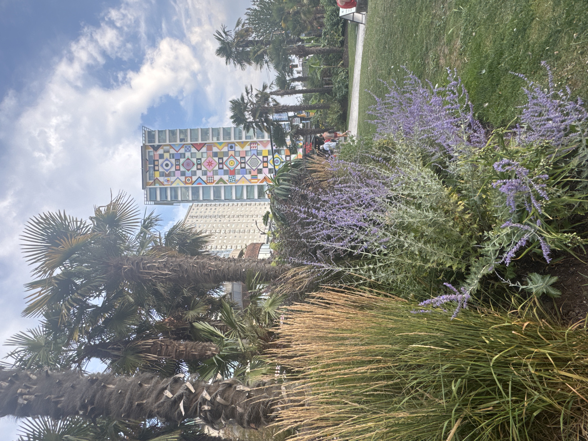 The image depicts the vibrant exterior of a building adorned with a colorful geometric mural, located in Vancouver, Canada. The mural is a striking visual element on the facade of a tall, modern building, featuring a mosaic of squares, circles, and diamond shapes in various bright colors. In the foreground, lush greenery includes tall, dry grass-like plants and flowering lavender bushes, suggesting a park or garden setting. Several palm trees with their distinctive fronds are also visible, contributing to a somewhat tropical ambiance within the urban landscape. In the middle ground, a paved pathway winds through the park, with a few individuals casually walking. The sky above is partially cloudy, with soft white clouds against a light blue backdrop. The overall scene conveys a lively and aesthetically pleasing urban environment with a strong artistic presence.
