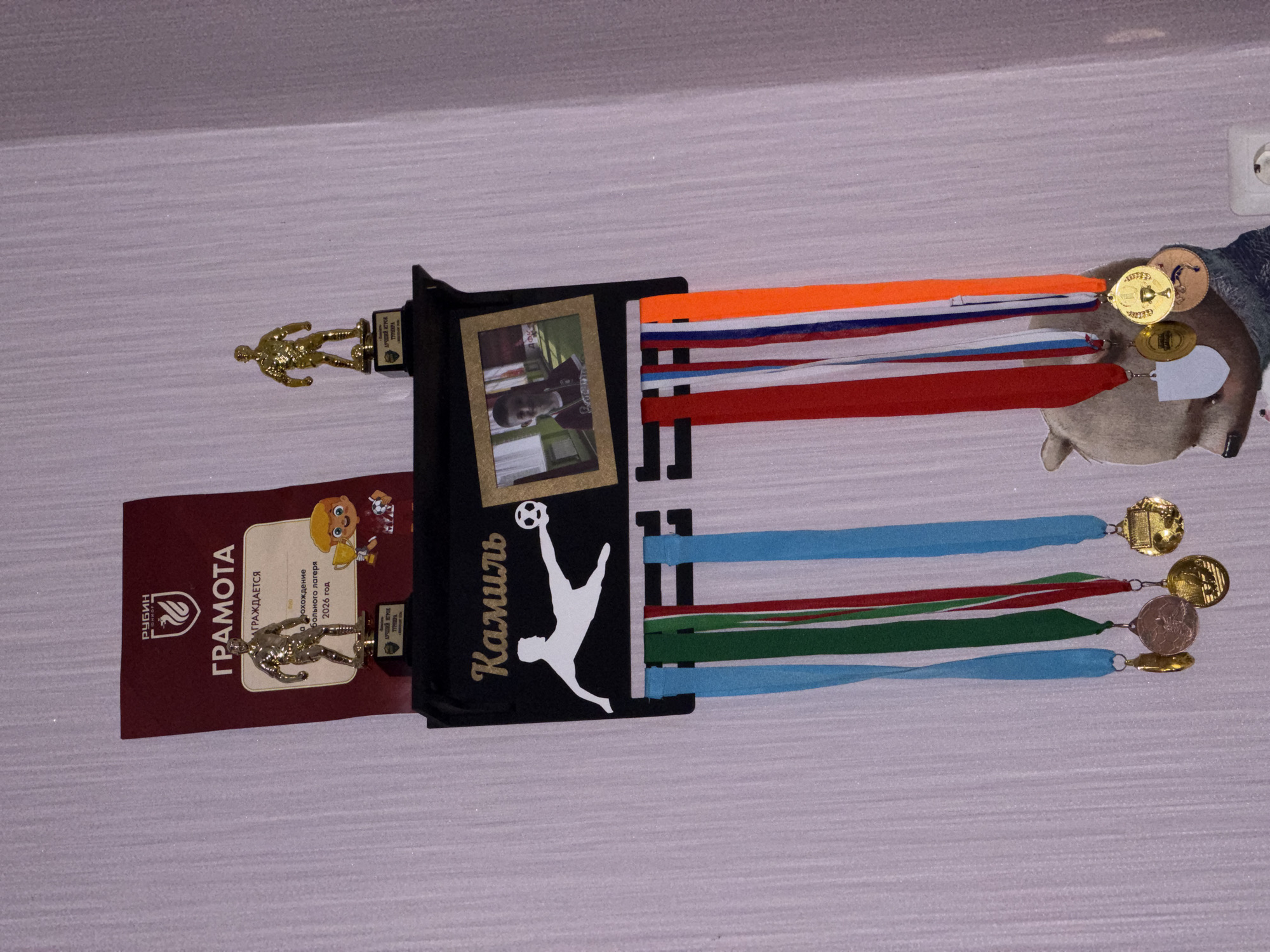 This image depicts a wall-mounted display of sports awards and memorabilia. Prominently featured is a dark shelf with the name "Камиль" (Kamil) written in a cursive font, accompanied by a silhouette of a soccer player in action and a framed photograph of a young boy. Below the shelf, several colorful ribbons hang, each attached to a medal. The ribbons are blue, green, red, white, and orange, suggesting participation in different events or teams. The medals themselves are varied in design, with some being circular and others shield-shaped, and appear to be made of gold and bronze-colored metal.
Above the shelf, a burgundy certificate with the "РУБИН" (Rubin) logo is partially visible, indicating an award. Two small golden trophies, one of a soccer player and another of a figure holding a cup, are placed on top of the certificate. A cartoon drawing of a child holding a trophy and a soccer ball is also present. The background is a light-colored, textured wall, possibly wallpapered, with a visible electrical outlet on the right side. The overall presentation suggests a dedicated space for showcasing achievements, likely in a home environment within the city of Tenishevo, Russia, though no specific landmarks or environmental features confirm the exact location.