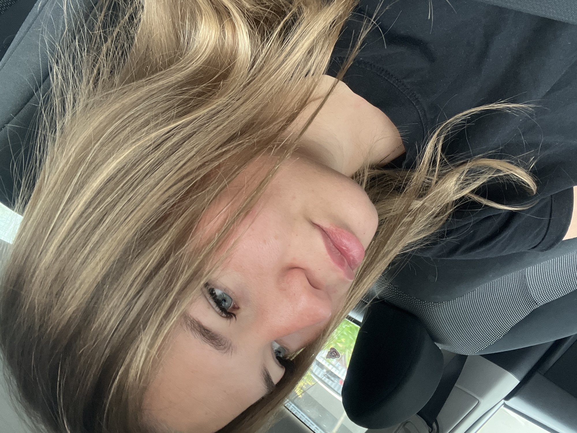 A young woman with light brown hair and blue eyes is in the driver's seat of a car. She is wearing a black shirt. The car interior is gray and black. The location may be Budva, Montenegro, but there is not enough visual evidence in the image to confirm the specific location. There is foliage visible through the car window.