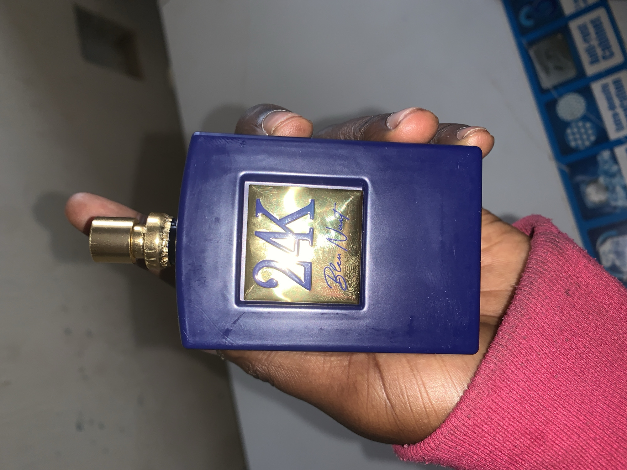 A close-up, eye-level shot captures a person's hand holding a dark blue rectangular bottle, likely a perfume or cologne, in an indoor setting. The hand, exhibiting a dark skin tone, holds the bottle horizontally with the thumb positioned on the left side near the gold-colored sprayer mechanism. Parts of the fingers are visible across the top and bottom of the bottle.
The bottle features a prominent gold rectangular label on its front, which displays the large blue text "24K" in a stylized font. Below "24K", a smaller script inscription reads "Bleu Night". The person's wrist or lower arm is partially visible on the bottom right, covered by a textured, pink fabric garment.
The background is out of focus. On the left, a plain, light-colored wall is visible. To the right, behind the hand, there's a blue and white patterned object, possibly packaging for another product, with some indistinct English text partially visible, including phrases like "fill-rest Cabinet" and "extra-durability." The lighting suggests an indoor environment. No specific activity or emotion is discernible other than the act of holding the bottle. This scene is provided with the location context of New Stadium, Nigeria.