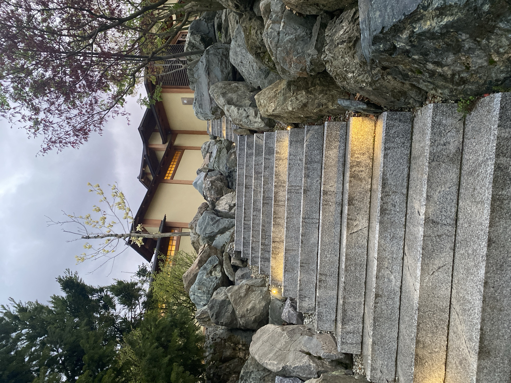 The image depicts a set of stone steps leading upwards alongside large rocks and vegetation. The steps are made of light gray, textured stone. Small lights are positioned between some of the steps, illuminating the path. Large, gray rocks border the steps on both sides, creating a natural, rugged feel. Green trees and branches with red and yellow leaves are visible on either side and above the steps. A building with a traditional Asian-style roof is partially visible in the background, with illuminated windows. The sky is overcast. The location is Krasnodar, Russia.