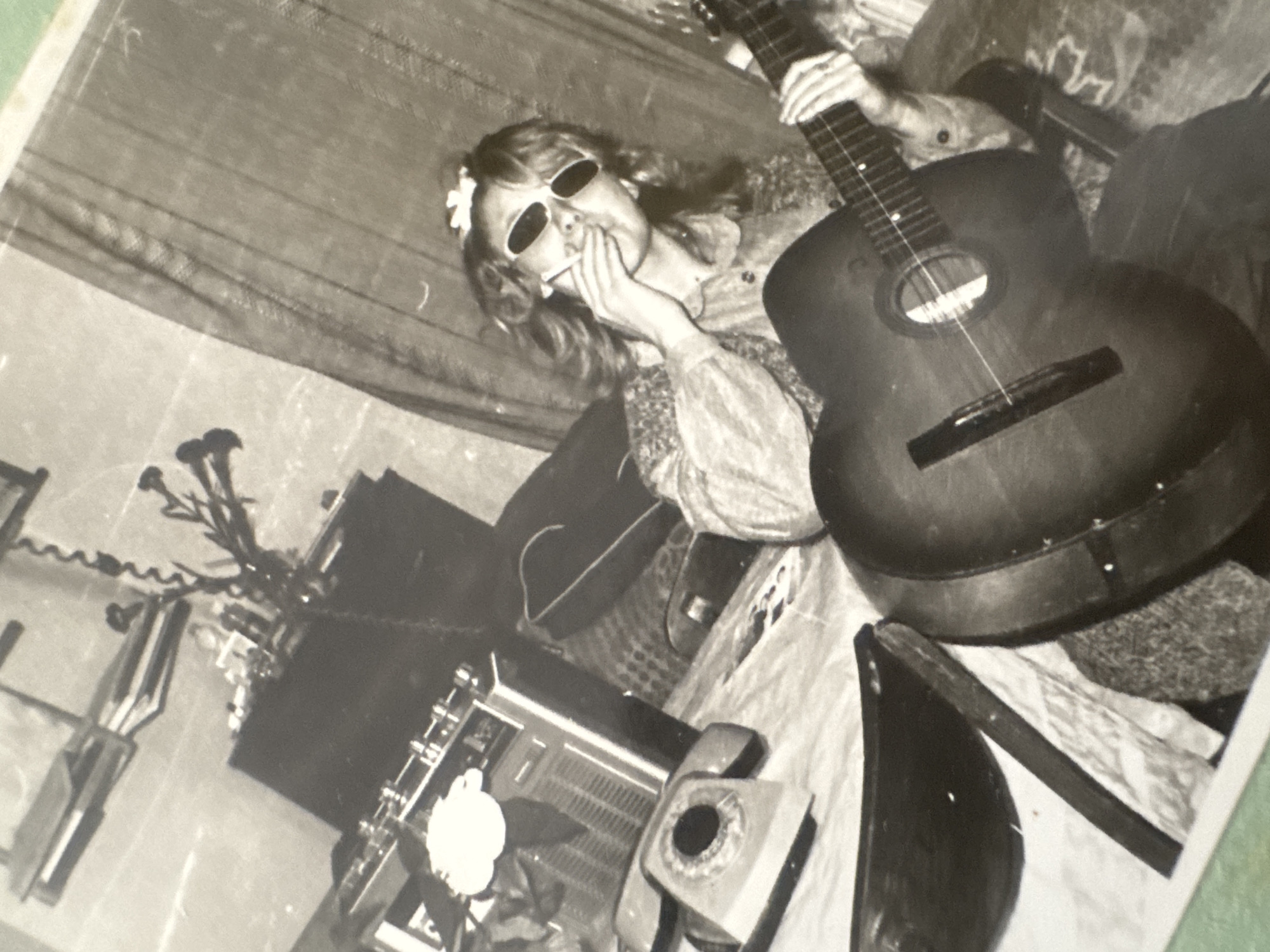 A black and white photograph shows a woman wearing dark sunglasses and a flower in her hair, holding a lit cigarette to her mouth. She has her other hand to her cheek. She is wearing a patterned blouse with puffy sleeves. The woman is sitting with an acoustic guitar in her lap. In the background is what appears to be a speaker, a telephone, a microphone stand with flowers in it and a partial view of a curtain. Based on the historical context of the image and the limited information available, the location in Ramat Gan, Israel cannot be determined from the image alone.