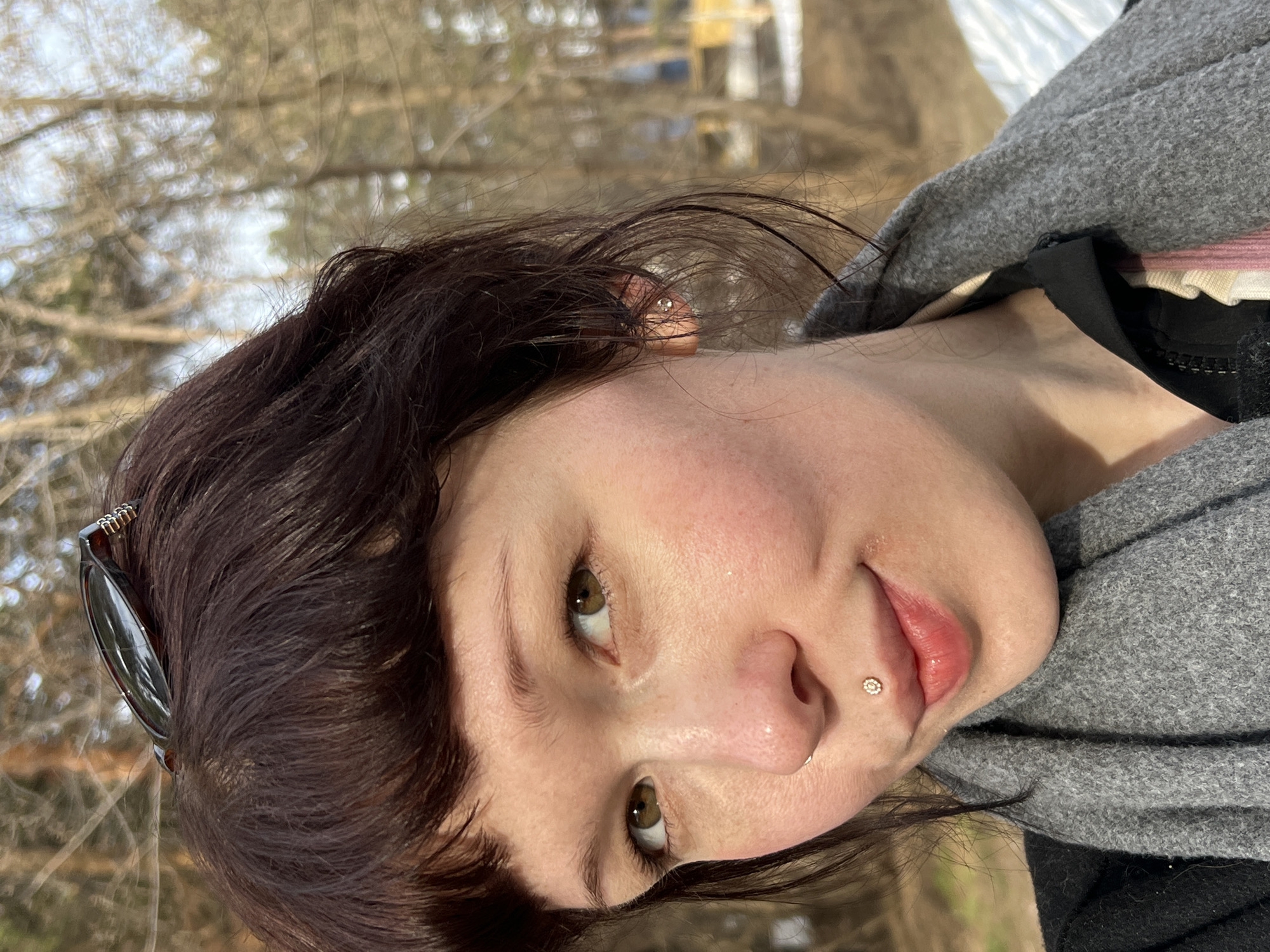 A close-up portrait shows a woman with dark brown hair and brown eyes. She has a nose piercing and a lip piercing. The woman is wearing a gray hooded sweater and a black top. Part of a hair clip is visible in her hair. The background features trees and a building. Given the location information (Chaikovsky, Russia), the landscape suggests an outdoor setting in Russia.