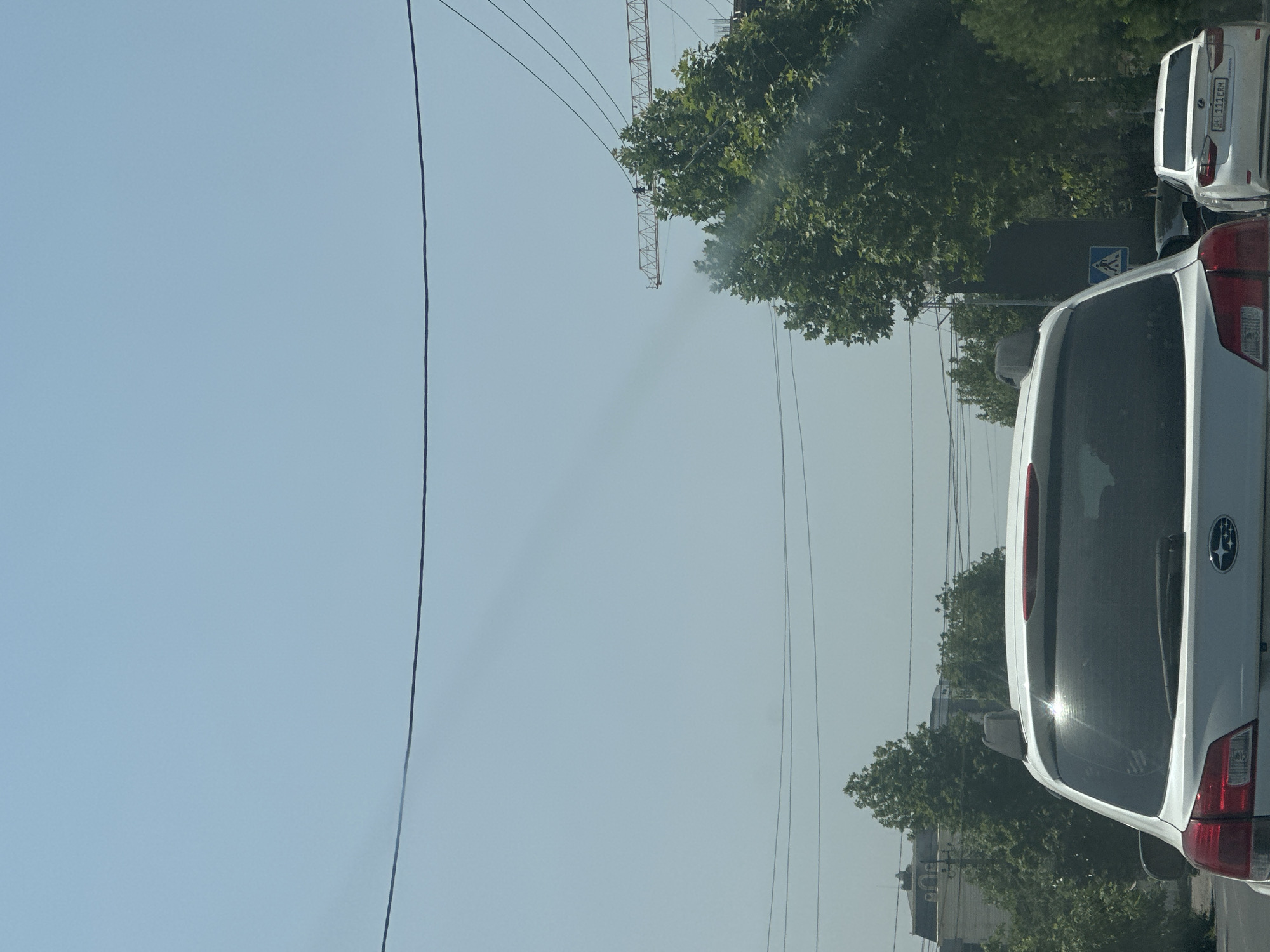 The image depicts a sunny daytime scene in Manas, Kyrgyzstan. In the foreground, the rear of a white Subaru Outback is visible, with its roof rack and spoiler prominently featured. To the right, a white Lexus sedan with a license plate reading "111 ERM" is parked. The background is dominated by lush green trees and a clear blue sky, with power lines crisscrossing the view. A construction crane is partially visible in the distance, suggesting ongoing development in the area. The image appears to have been taken from a vehicle, likely in traffic, given the perspective and the presence of other cars.