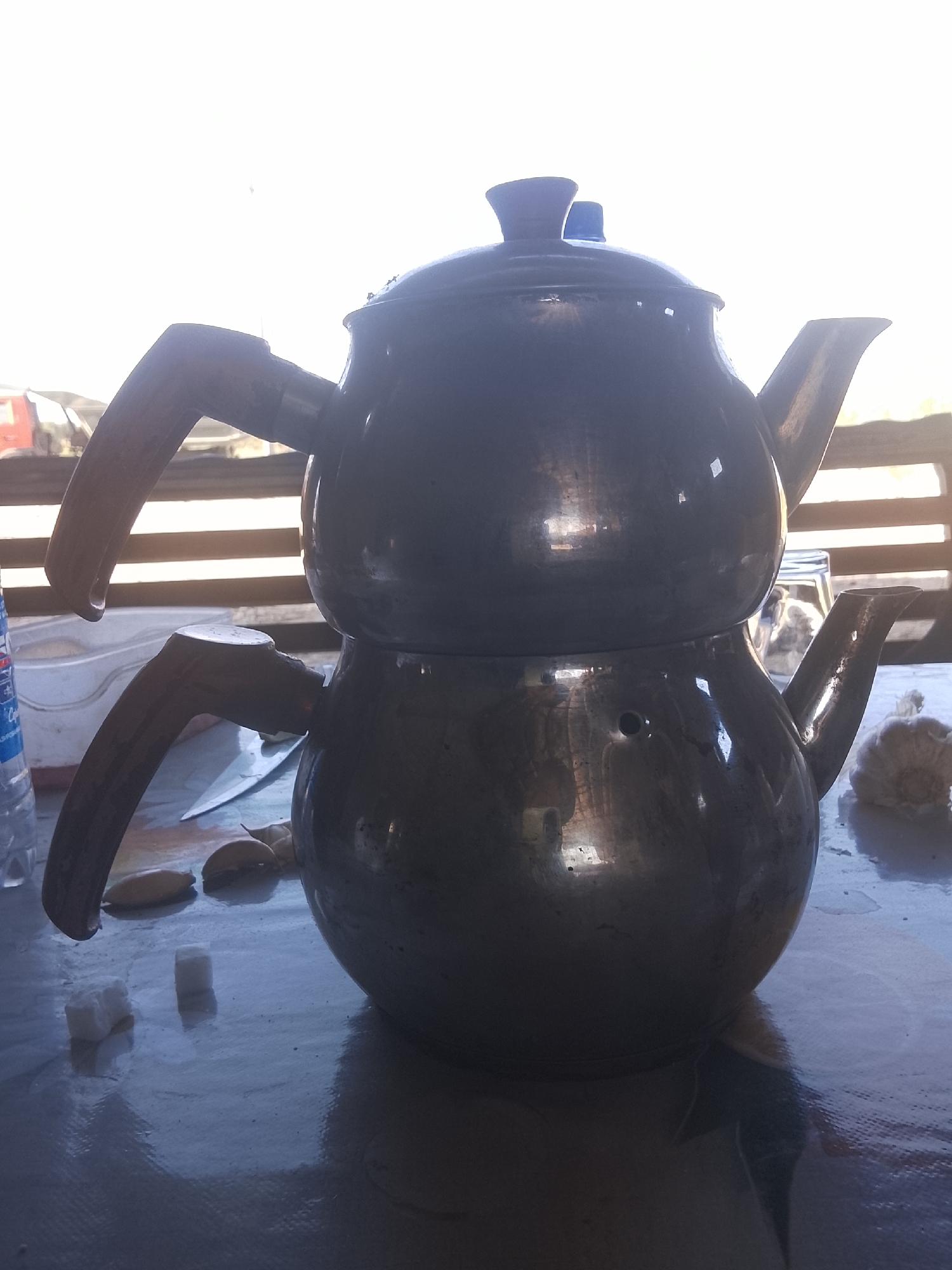 An outdoor setting, possibly a balcony or patio, bathed in bright sunlight with a blurry, light-colored background. In the foreground, a dark, metallic two-tiered teapot with wooden handles sits on a textured surface, likely a table. A blue plastic water bottle, a white plastic container, and a couple of sugar cubes are visible to the left of the teapot. To the right, a light-colored, amorphous object, possibly food or a decorative item, is present. The overall impression is that of a casual, domestic scene. The location is identified as Sabir Rakhimov, Uzbekistan.