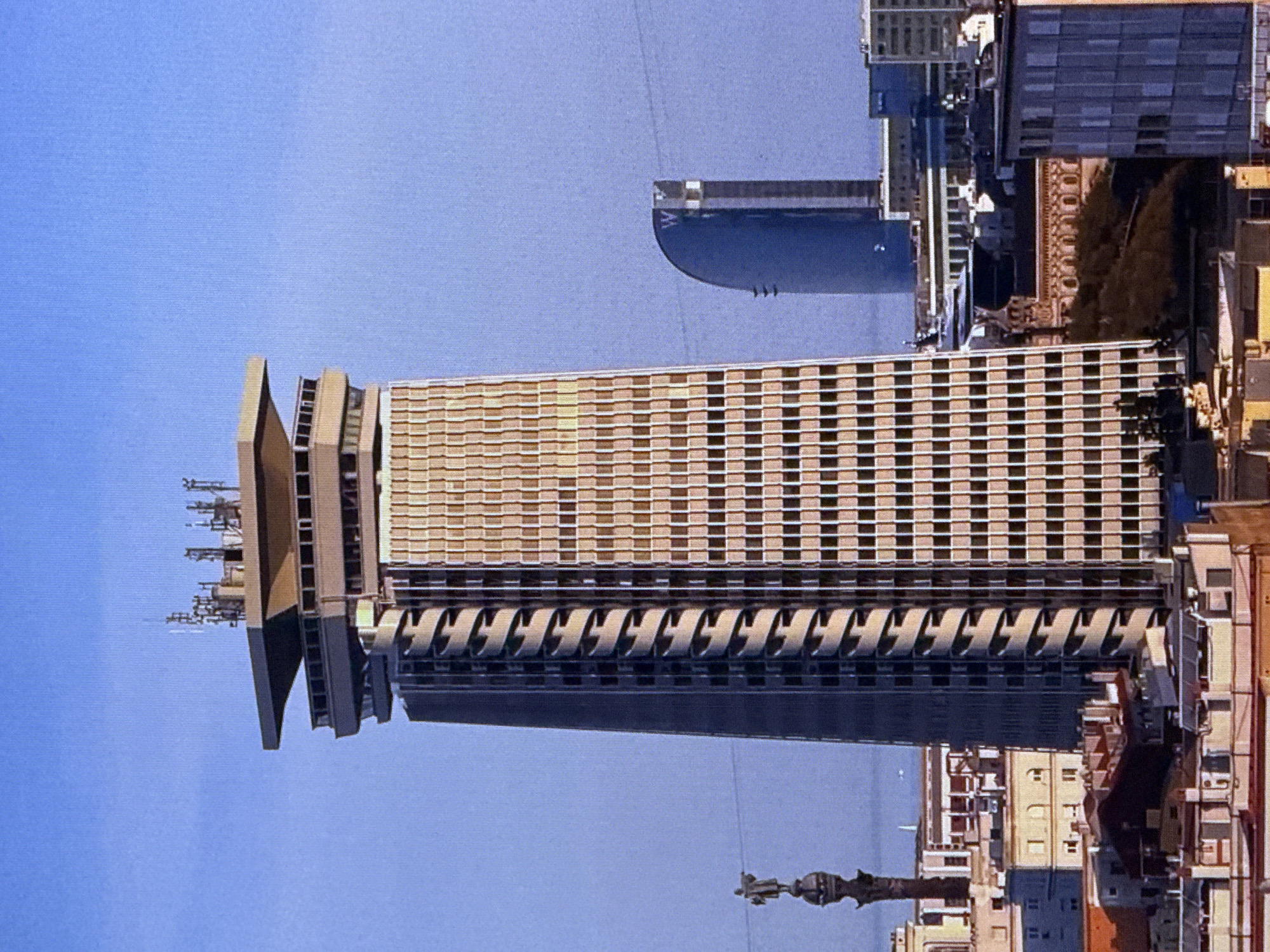 The image displays a cityscape dominated by the Edificio Colón, a tall, distinctive skyscraper in Barcelona, Spain, characterized by its cylindrical spiral facade. In the background, the curved, sail-like form of the Hotel W Barcelona is visible, further indicating the Barcelona location. To the left of the Edificio Colón, a statue atop a column, likely the Columbus Monument, stands against a clear blue sky. The foreground and middle ground are filled with various buildings, showcasing a mix of architectural styles typical of a European urban environment. The scene is static, with no visible people or discernible actions, offering a panoramic view of the city's architecture under daylight.