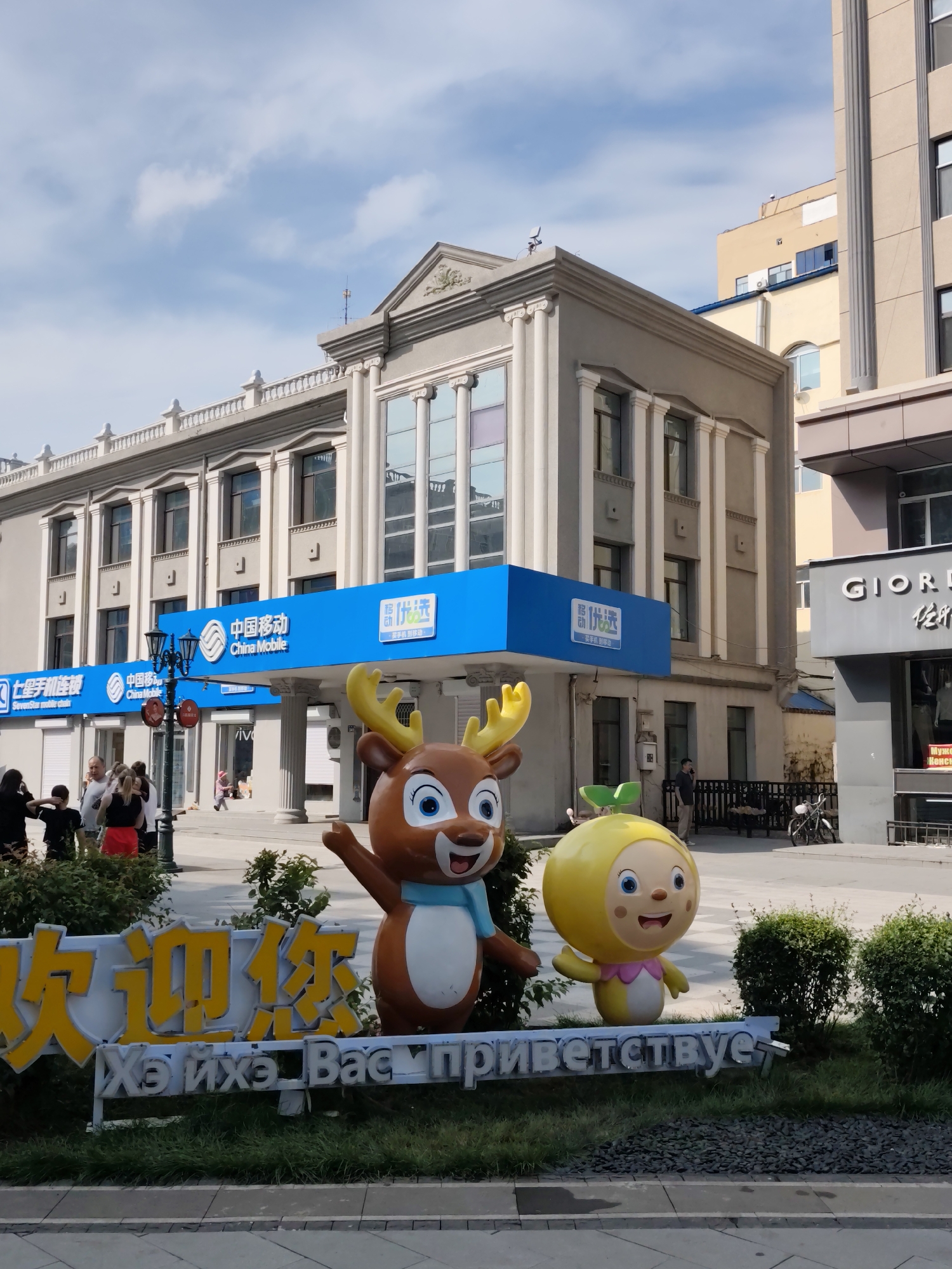 In Aihui, China, a scene unfolds under a partly cloudy sky, featuring a building with a classical facade, adorned with columns and a pediment. The building's exterior is painted a light gray, and it houses multiple businesses, including "China Mobile" prominently displayed on a bright blue sign. To the right of the main building, a section of a beige building is visible, and further right, a gray facade with "GIORD" in large letters suggests another commercial establishment.
In the foreground, two large, colorful cartoon-like statues stand guard. The statue on the left depicts a brown deer with large antlers and a blue scarf, its right arm raised in a waving gesture. The statue on the right is a yellow, spherical character with arms and legs, a green sprout on its head, and a pink bow around its neck, also with its right arm raised.
In front of these statues, a sign with yellow Chinese characters and white Cyrillic lettering that reads "Хэихэ Вас приветствует" (likely meaning "Heihe Welcomes You") is partially obscured by grass and gravel.
Several people are present in the mid-ground, some walking, others standing and conversing. They are casually dressed, indicating a typical day in a public urban space. A black lamppost stands near the entrance of the building, and a bicycle is parked near the beige building. The ground is paved with gray tiles, and manicured bushes are scattered throughout the area, contributing to the urban landscaping.