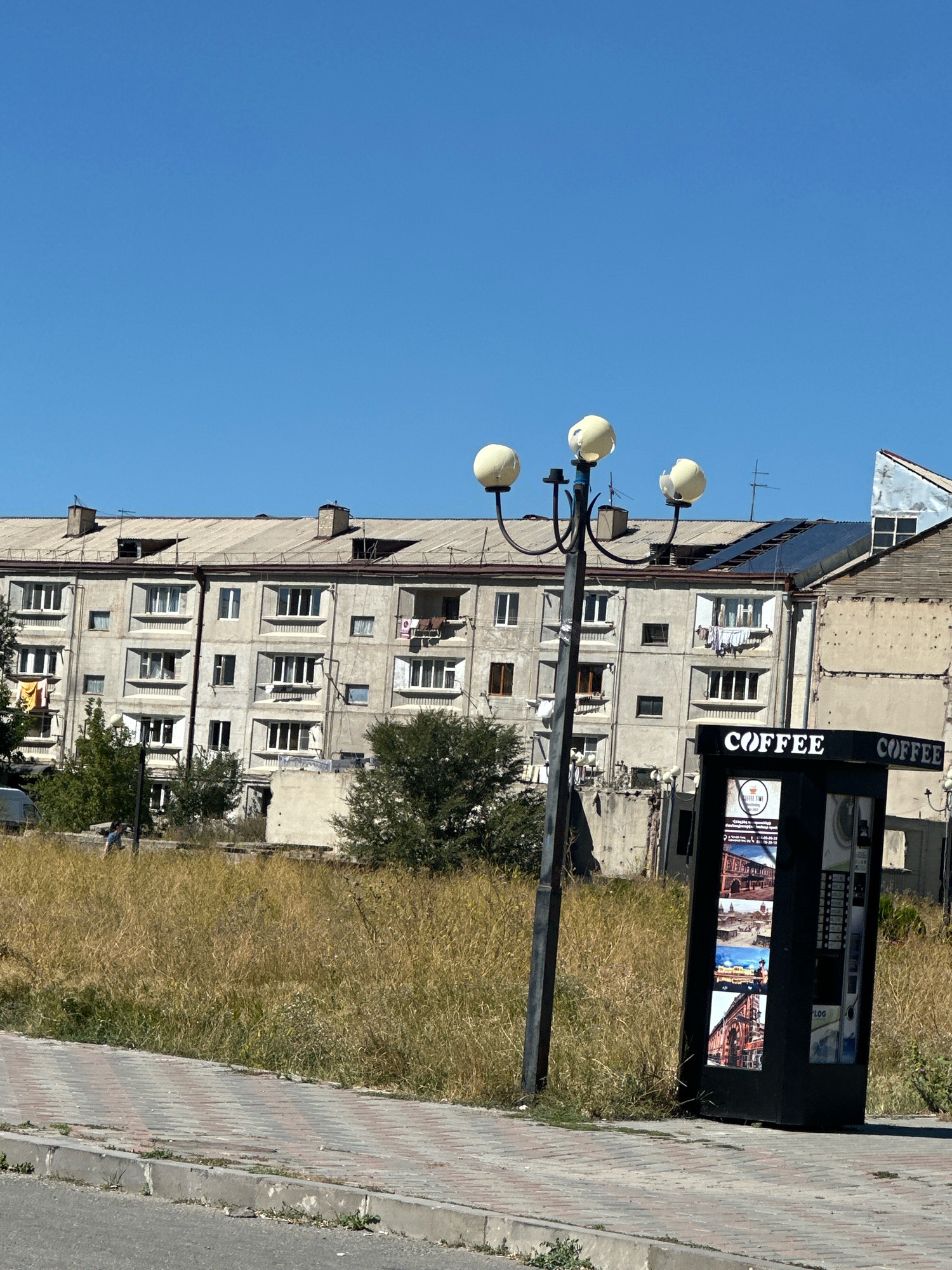 The image shows a street scene in the city of Gyumri, Armenia. In the foreground, a paved sidewalk runs alongside a street. To the right of the sidewalk is a black coffee vending machine. Behind the sidewalk, a patch of dry, overgrown grass is visible. In the background, a large, multi-story apartment building with a grey facade and numerous windows stands under a clear blue sky. A street lamp with three spherical lights is positioned in front of the building and the vending machine. The architecture of the building suggests it is an older Soviet-era structure common in Gyumri. The overall impression is one of an urban environment with signs of wear and tear, interspersed with modern amenities like the coffee machine.