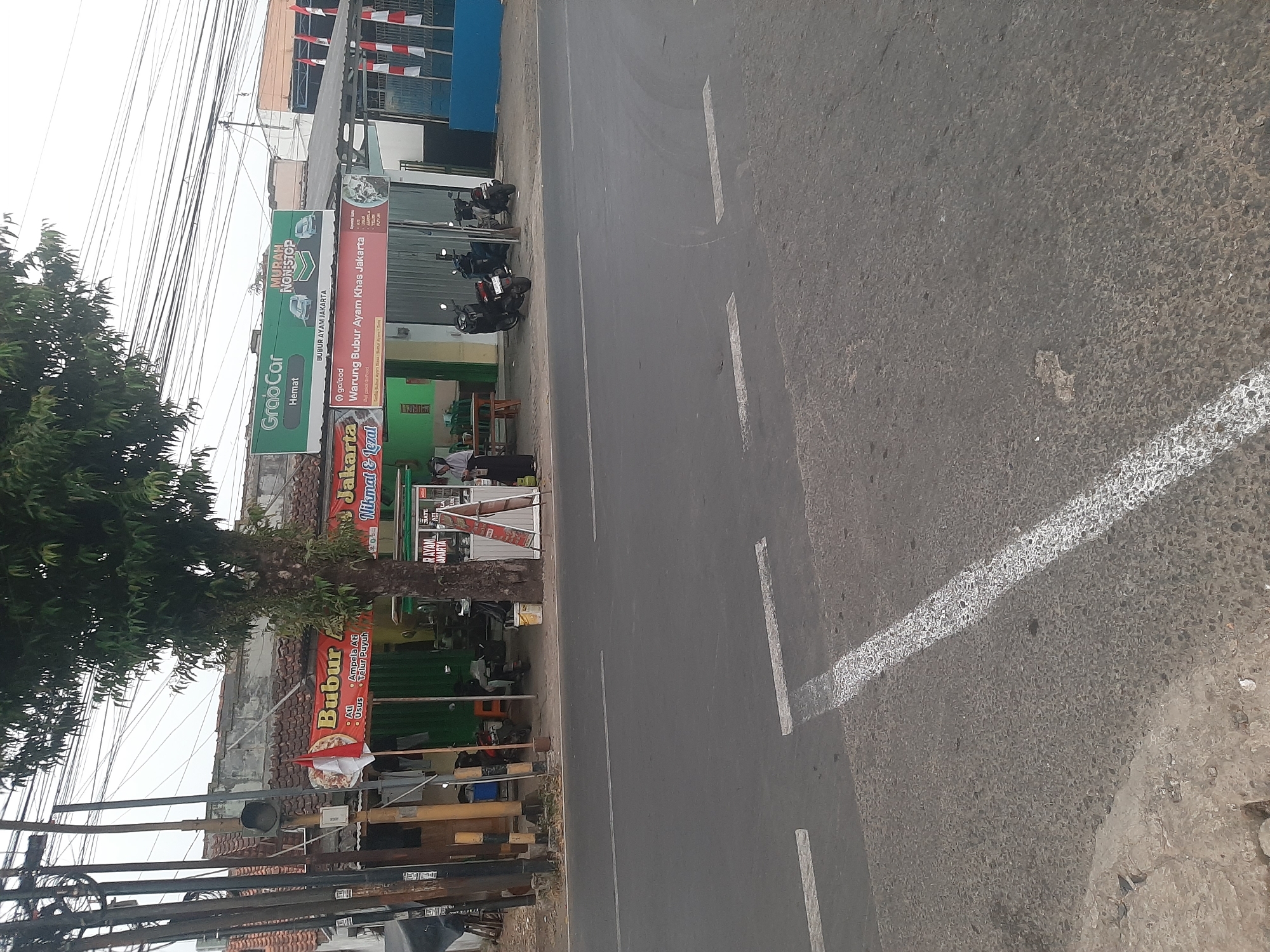 In the city of Purwanegara, Indonesia, a street scene unfolds with a focus on a small food establishment and its surroundings. The image captures a building with a tiled roof, partially obscured by a large tree. A red banner on the building reads "BUBUR AYAM JAKARTA" and "Nikmat & Lezat," indicating it is a chicken porridge restaurant. Next to it, a sign for "GrabCar Hemat" and "MURAH NON-STOP" suggests the presence of ride-sharing services. A woman in a hijab stands near a food cart, and several motorcycles are parked in front of the establishment. The street in the foreground is paved and shows white lane markings. Electrical poles and wires are visible along the side of the road, typical of urban Indonesian infrastructure. The scene appears to be during daylight, with an overcast sky.