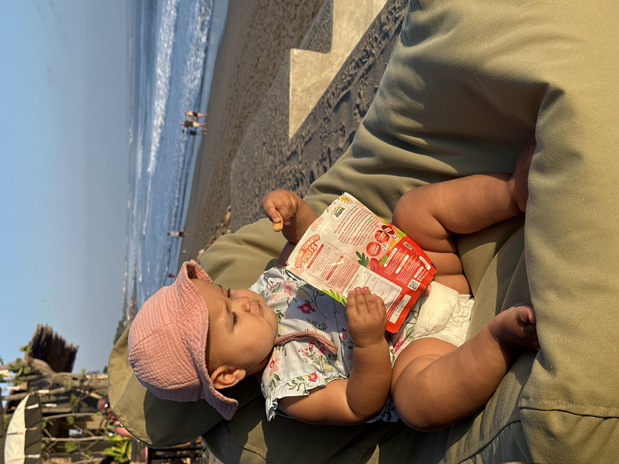 A toddler wearing a pink sun hat and a floral print dress sits in a large beanbag chair on a sandy beach. The toddler is holding a red snack packet and a single snack in their right hand. The ocean and waves are visible in the background, with several people walking along the shore. To the left of the toddler, a thatched-roof structure with umbrellas is partially visible, suggesting a beachfront establishment in North Kuta, Indonesia. The sun is low in the sky, casting long shadows and bathing the scene in warm, golden light.