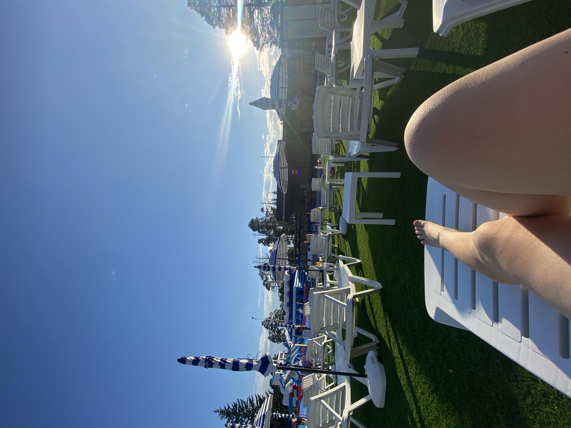 This media file depicts a bright, sunny day at an outdoor water park in Votkinsk, Russia. The scene is set on a grassy area, likely artificial turf, with numerous white lounge chairs and tables arranged for visitors. In the foreground, a person's legs and feet are visible, resting on one of the white lounge chairs, suggesting a relaxed atmosphere and enjoyment of the sunny weather.
In the background, a water park with inflatable slides and structures, primarily in blue and white, is visible. Several striped umbrellas, also in blue and white, are set up around the lounge area, providing shade. The sun is shining brightly in the sky, creating lens flare and casting shadows on the ground, indicating it is likely midday. The clear blue sky suggests fair weather. The presence of water park attractions and lounge furniture clearly points to a recreational setting. The overall mood conveyed is one of leisure and enjoyment on a warm summer day. No discernible text is visible in the image.