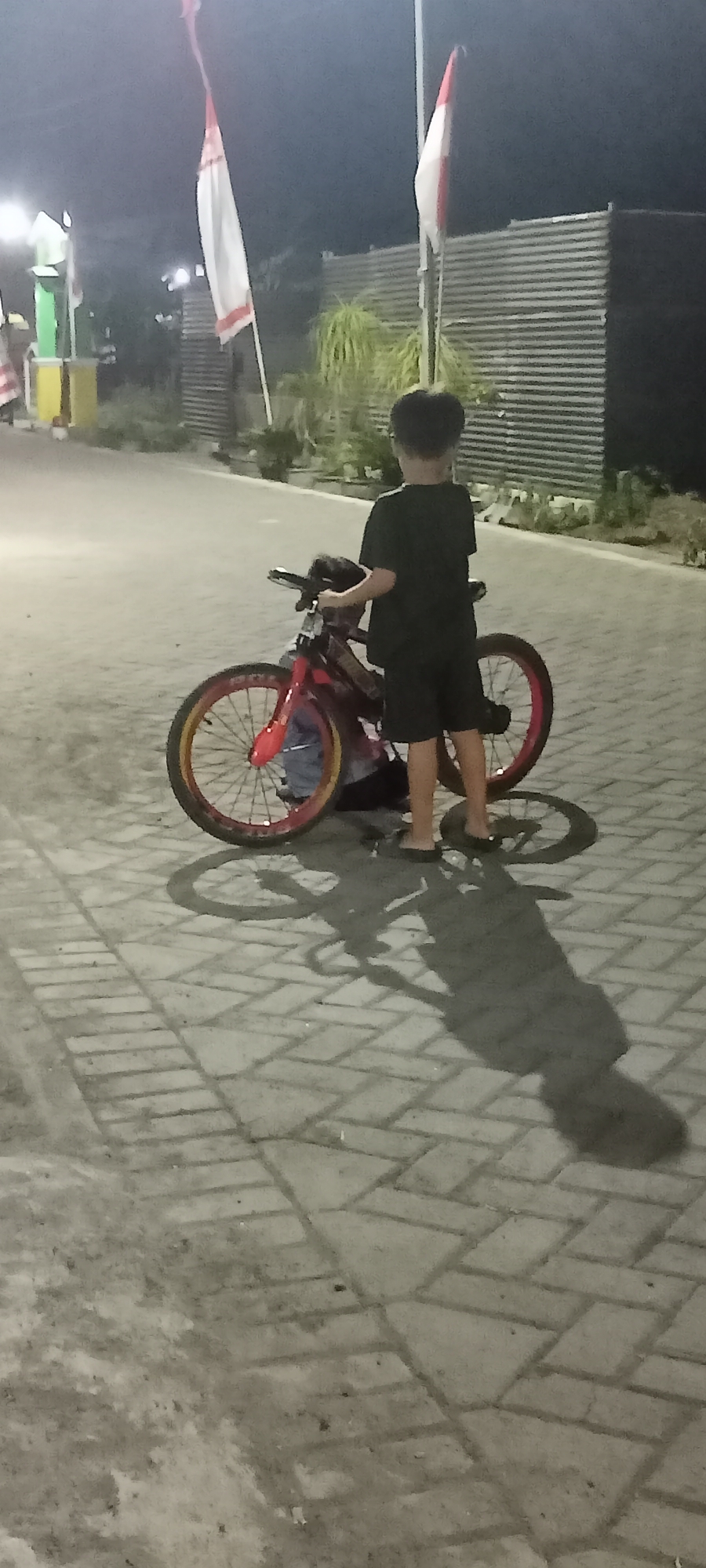 In Gempolklutuk, Indonesia, at night, a young boy stands beside a red bicycle on a paved walkway. The boy, wearing a black t-shirt and shorts, holds the handlebars of the bicycle, which has red and yellow rims. His shadow stretches across the patterned grey pavement. In the background, two Indonesian flags, red and white, fly from poles. A corrugated metal structure and some green foliage are also visible. The lighting suggests artificial illumination, likely from streetlights.