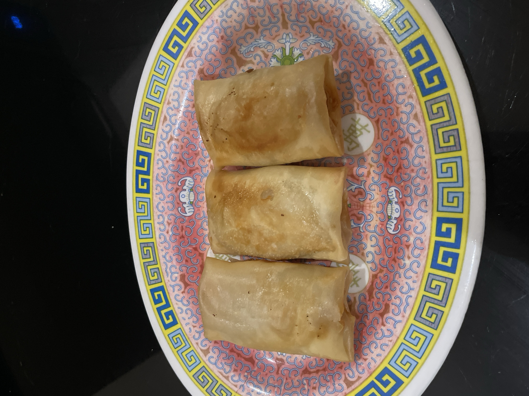 A top-down shot shows three golden-brown spring rolls arranged vertically on a decorative plate. The plate has a white base with a pink and blue swirling pattern and a yellow and blue geometric border. The spring rolls are cylindrical and appear to be lightly fried, with some subtle browning on their surfaces. They are positioned close together, occupying the center of the plate. The background is a dark, out-of-focus surface, likely a table. The lighting is direct, highlighting the texture and color of the spring rolls and the details of the plate. There are no people visible in the image, and no specific time of day, weather, or location cues are evident. There is no visible text in the image.